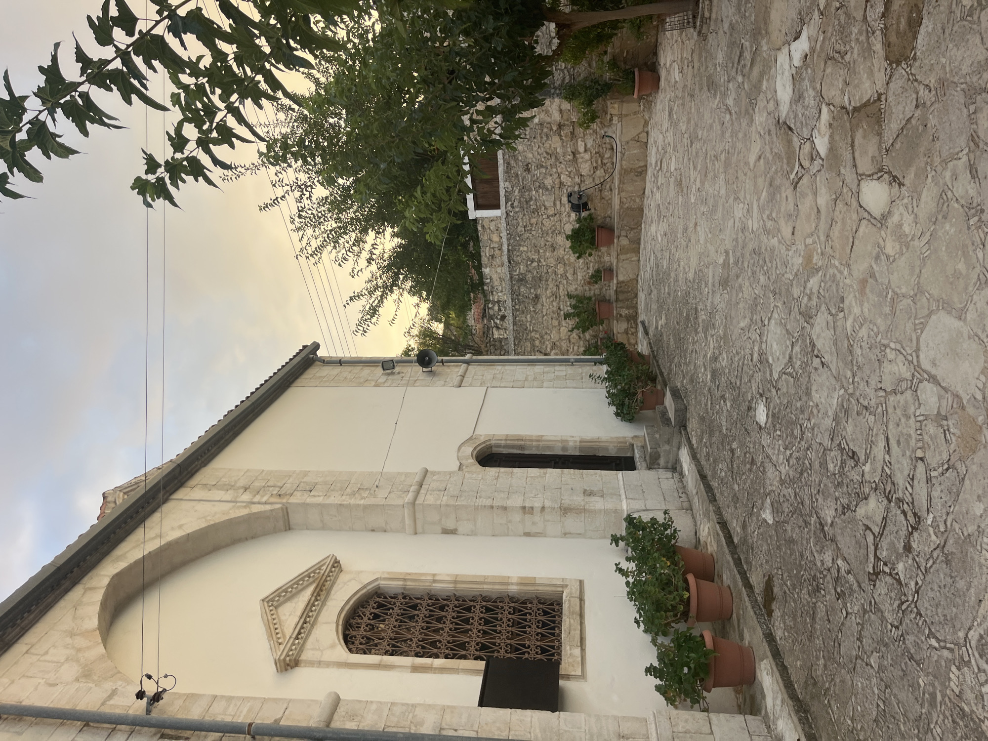 The image depicts a view of a courtyard in Lofou, Cyprus, featuring a white-washed building with stone accents and arched architectural elements. A prominent arched window with intricate metalwork is visible on the left side of the building, adorned with a triangular stone pediment above. Adjacent to this window is a dark, arched doorway. Several terracotta pots with green plants are arranged along the building's base and the courtyard's edge. The courtyard itself is paved with irregular, light-colored flagstones interspersed with darker grout lines.
In the background, a stone wall separates the courtyard from a more distant area with trees and a wooden-doored structure. A large tree with dark green foliage extends into the frame from the right, its branches reaching towards the sky. The sky above is overcast with soft, warm light, suggesting either dawn or dusk. Electrical wires are strung across the sky, supported by poles visible on the left side of the building. The overall scene conveys a sense of peaceful, traditional architecture in a Mediterranean setting.