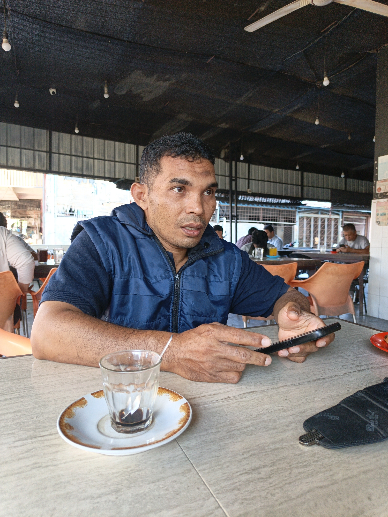 An adult man sits at a table in an indoor public space, possibly a restaurant or cafe, in Lhokseumawe, Indonesia. He is wearing a dark blue short-sleeved shirt under a blue vest. He holds a black smartphone in his hands, looking towards the right of the frame.
On the table are a small, clear glass and saucer containing the remnants of a dark liquid, and a black object that appears to be a sunglasses case. The table is light-colored and appears to have a tiled surface.
In the background, other people are seated at tables, also eating or drinking. The space has a partially open structure, with corrugated metal roofing and supporting beams visible. The lighting is provided by hanging bulbs. The chairs are orange plastic. The scene suggests a casual, daytime setting.