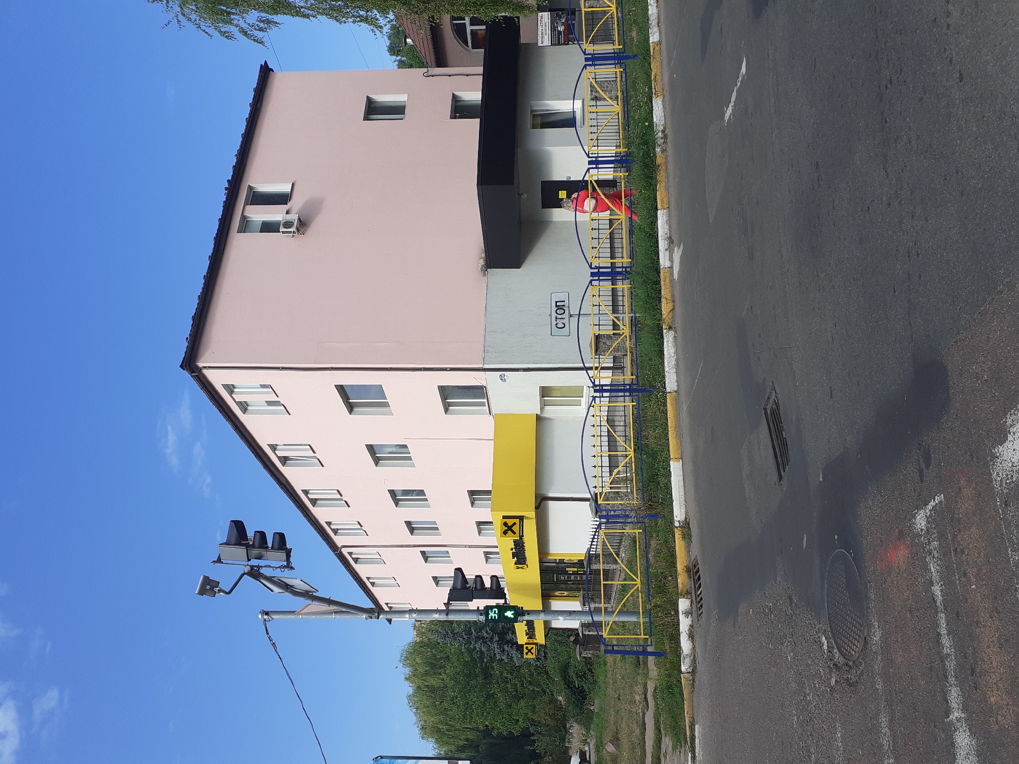 The image depicts a street scene in Obukhiv, Ukraine, on a clear, sunny day. In the foreground, a paved road with white lane markings and a manhole cover is visible. Adjacent to the road is a patch of overgrown grass bordered by a yellow and blue fence. Behind the fence stands a light pink, multi-story building with several windows.
A yellow and white structure with the "Raiffeisen" logo is attached to the front of the pink building. A traffic light with a pedestrian signal showing a green walking figure and the number "35" is positioned on the left side of the scene, near the Raiffeisen building. To the right of the Raiffeisen building, a smaller white section of the building has a black overhang and a door. A sign with the Cyrillic word "СТОП" (STOP) is mounted on the wall above this section.
A woman wearing a bright orange tracksuit walks past the entrance of the building, carrying a white bag. In the background, trees and other buildings are visible. The sky is blue with scattered white clouds.