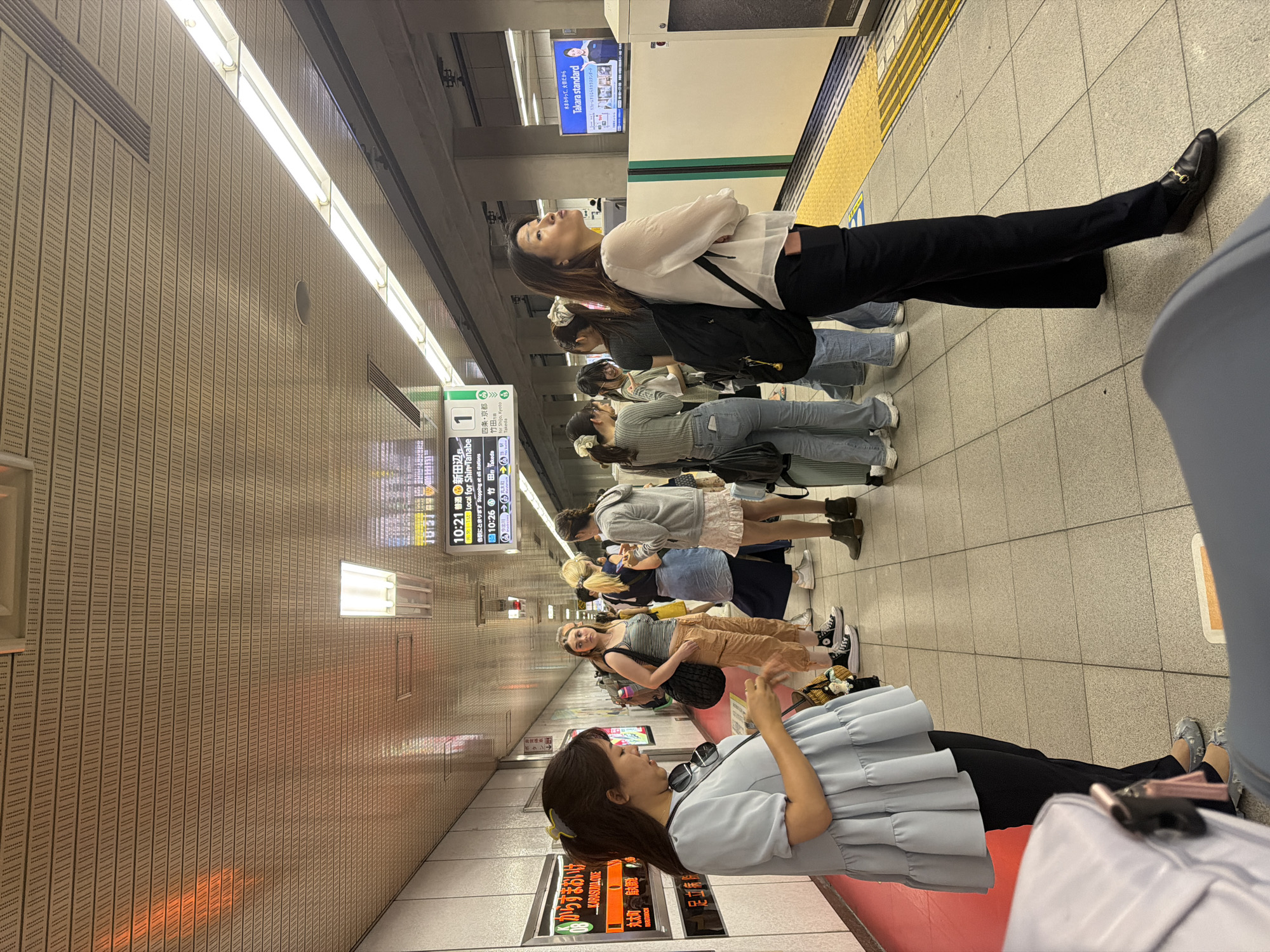 This is a medium shot of a group of people waiting on a subway platform in an indoor station, with a train track visible. The platform has a yellow safety line along the edge. The ceiling is made of beige acoustic tiles with a pattern of small holes. Overhead fluorescent lights illuminate the space. A digital sign displays train information, including times and destinations.
In the foreground, a woman with dark hair pulled back is looking down at her phone. She is wearing a light blue ruffled tunic and black pants. To her right, a young woman with light brown hair in a ponytail is wearing a grey and white striped tank top and beige cargo pants, holding a black bag. Behind her, a woman with blonde hair in a bun is wearing a grey hooded jacket over a white top and a light blue skirt. Further back, another group of people are standing, including a woman in a grey shirt and jeans, and a woman with a backpack. A train is partially visible on the tracks to the right. An advertisement for "Takara standard" is displayed on a blue and white billboard in the background. The floor is tiled in a grid pattern.