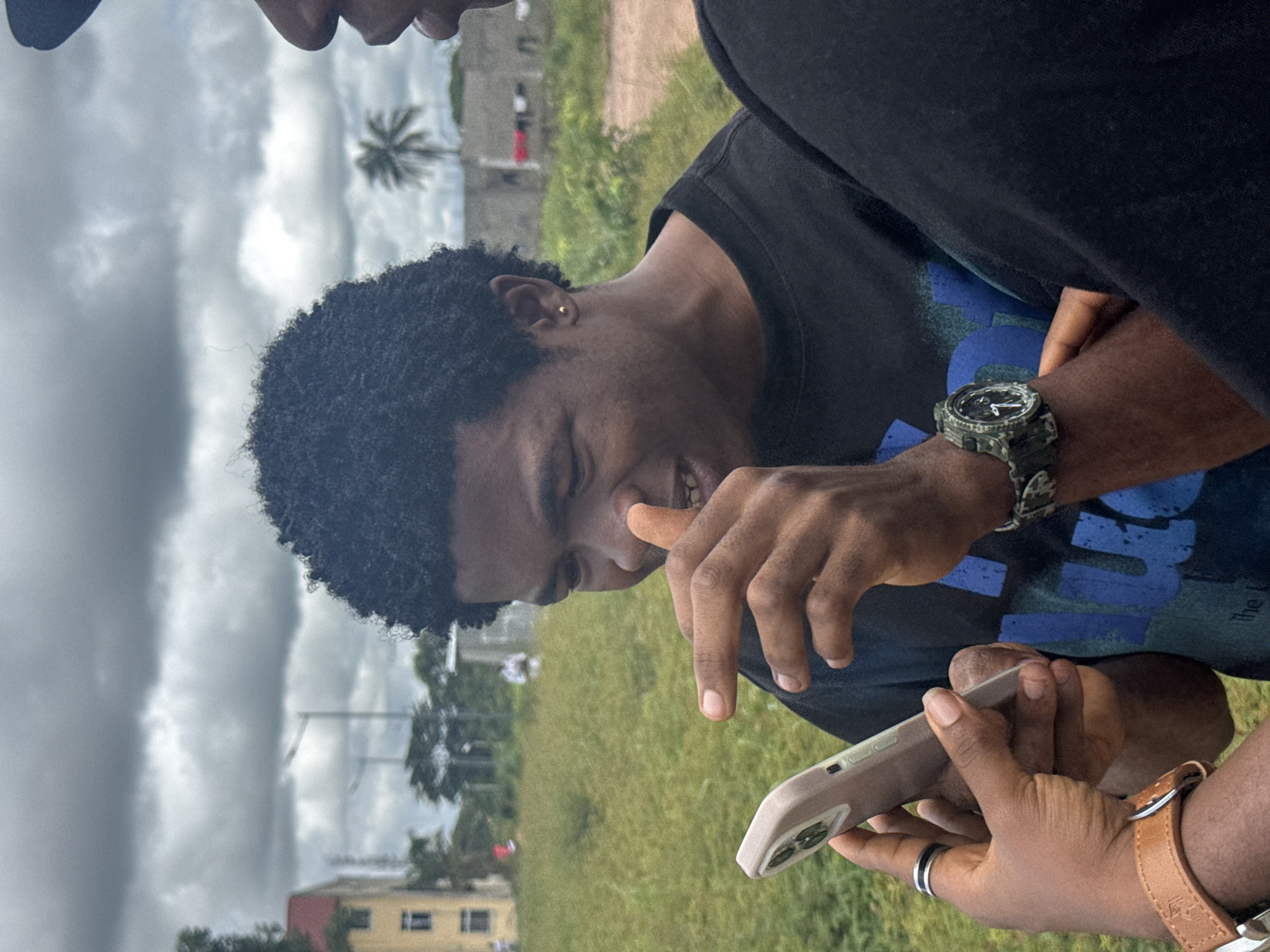 An outdoor medium shot captures two young Black men in Ihiagwa, Nigeria, as one, in the foreground, examines a smartphone held by another. The man in the foreground, with curly black hair and an earring, is looking down at the phone, his mouth slightly open, and his right index finger near his nose, as if in thought or contemplation. He wears a black t-shirt with a blue design. The man holding the phone, whose face is mostly out of frame to the right, has a darker complexion and is wearing a black baseball cap and a black shirt. He appears to be handing the phone to the man in the foreground, or has just handed it over. Both men have dark skin tones and are dressed casually. The man in the foreground wears a silver ring on his left index finger and a brown leather-strapped watch on his left wrist. The other man has a green camouflage watch on his right wrist.
The environment is outdoors with a cloudy sky. In the background, there is a grassy field with some sparse trees. To the left, a building with a red roof and yellow walls is visible. Further back, there are other structures, some appearing to be under construction or unfinished, with laundry hanging outside. The overall setting suggests a semi-urban or rural area within Ihiagwa. The lighting is diffused due to the overcast sky. The image seems to be taken during the daytime.