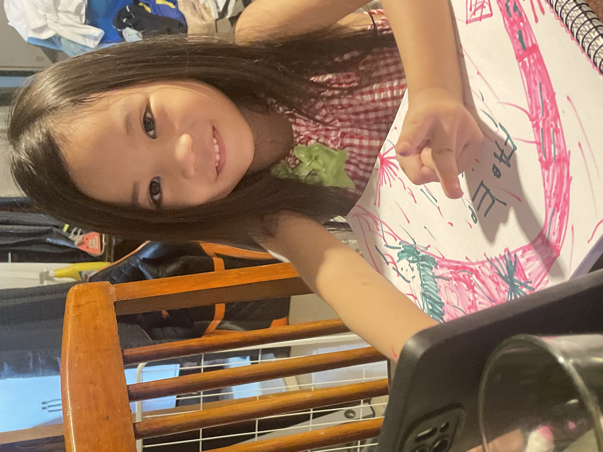 The image shows a young girl with long, dark hair and a red and white checkered dress. She is smiling and looking at the camera while holding a piece of paper with a drawing on it. The drawing appears to be done with markers, with bold pink and green lines. She is seated at a wooden table, with a phone in the foreground. The background is slightly out of focus but shows a domestic setting with some laundry and bags visible. The lighting suggests it might be daytime. The girl's expression conveys a sense of happiness and engagement with her artwork. The location context is Davao City, Philippines, but there are no specific visual cues in the image to confirm this.