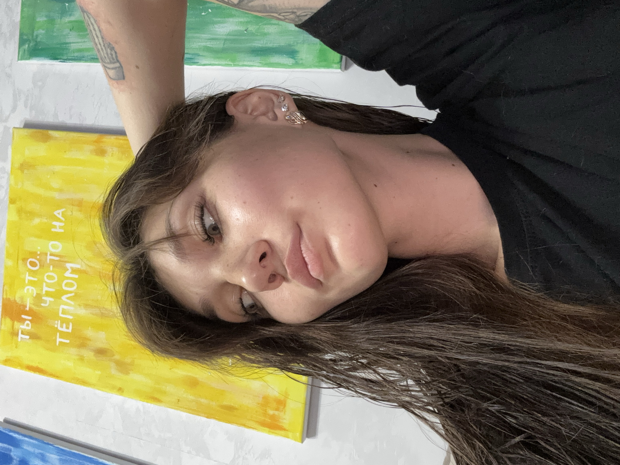 A young woman with long, dark, wet hair and light brown eyes is pictured from the chest up, against a wall with paintings. She is wearing a black t-shirt and has two earrings in her left ear. Her right arm is raised and resting on the back of her head, revealing a tattoo on her upper arm. She is looking off to her left. The paintings in the background are abstract. One is yellow with white text, and another is green with white stripes. The overall scene is indoors, with artificial lighting. The location is not specified in the image.