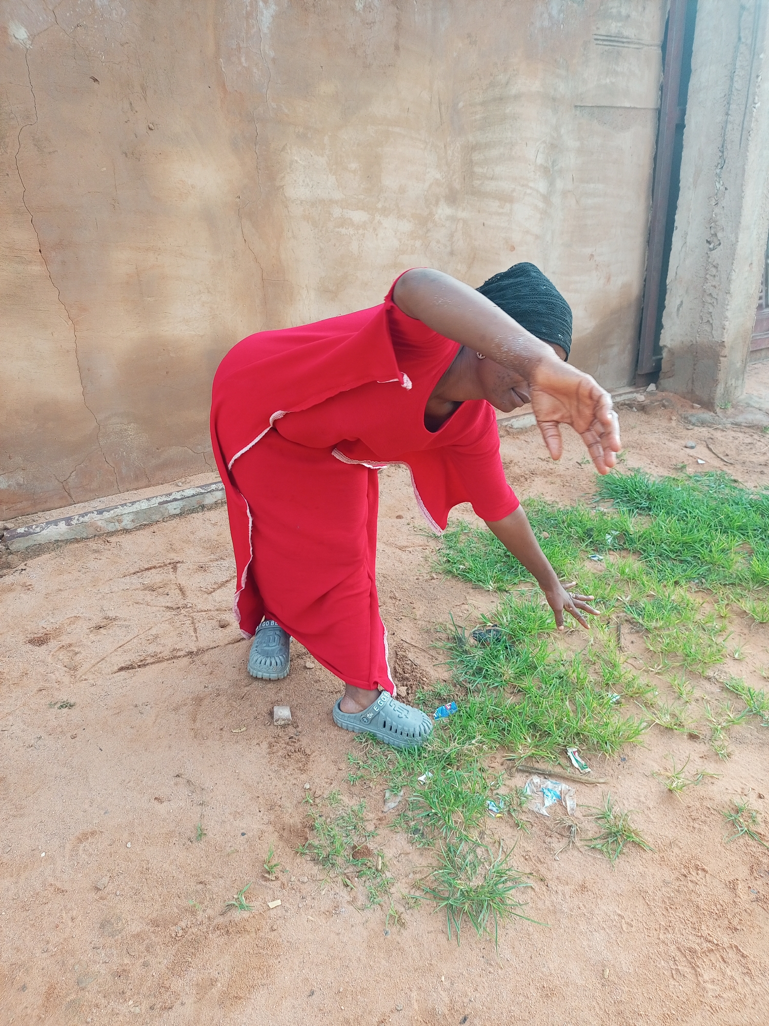 A Black woman wearing a red dress and a black headscarf is bending down to pick something from the ground. She is outdoors, in what appears to be an undeveloped area with a sandy ground and some patches of grass. The wall behind her is made of a light-colored, textured material, possibly concrete or stucco, with some cracks and water stains. To the right of the wall, there is a dark vertical post, possibly part of a building's structure or a gate. The lighting suggests it is daytime.