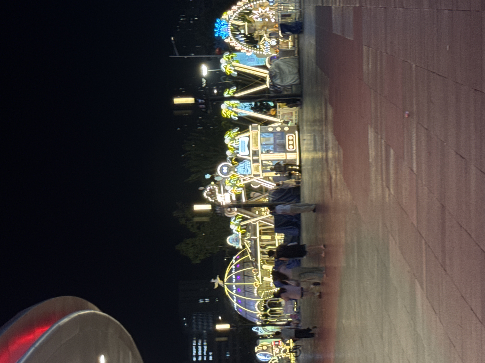 In Shenyang, China, at night, an outdoor plaza is illuminated by an array of vibrant decorative lights. Several people are present, casually walking across the paved area. In the background, a collection of illuminated structures resembling carnival rides or attractions are visible, including a dome-shaped structure, a bus-like structure, and a Ferris wheel, all adorned with numerous lights. Trees can be seen behind the attractions, and the outlines of buildings are discernible in the distance. The ground is paved with large, rectangular tiles, reflecting the ambient light. The overall scene suggests a festive or entertainment area open to the public.