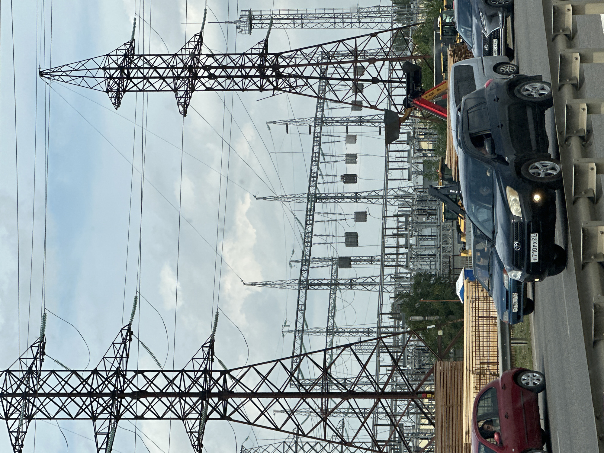 The image depicts a view of an industrial area with power lines and electrical substations, likely in or near the city of Beryozovy, Russia. In the foreground, several cars are driving on a road. A red hatchback is on the left, a blue sedan is in the middle, and a black SUV is on the right, followed by a silver minivan and a grey Lexus SUV. The vehicles are in motion, suggesting traffic. In the background, large power transmission towers and a complex electrical substation are visible. Construction equipment, including what appears to be a loader and stacked lumber, are also present. The sky is overcast with clouds. The scene appears to be a functional, active industrial and transportation corridor.