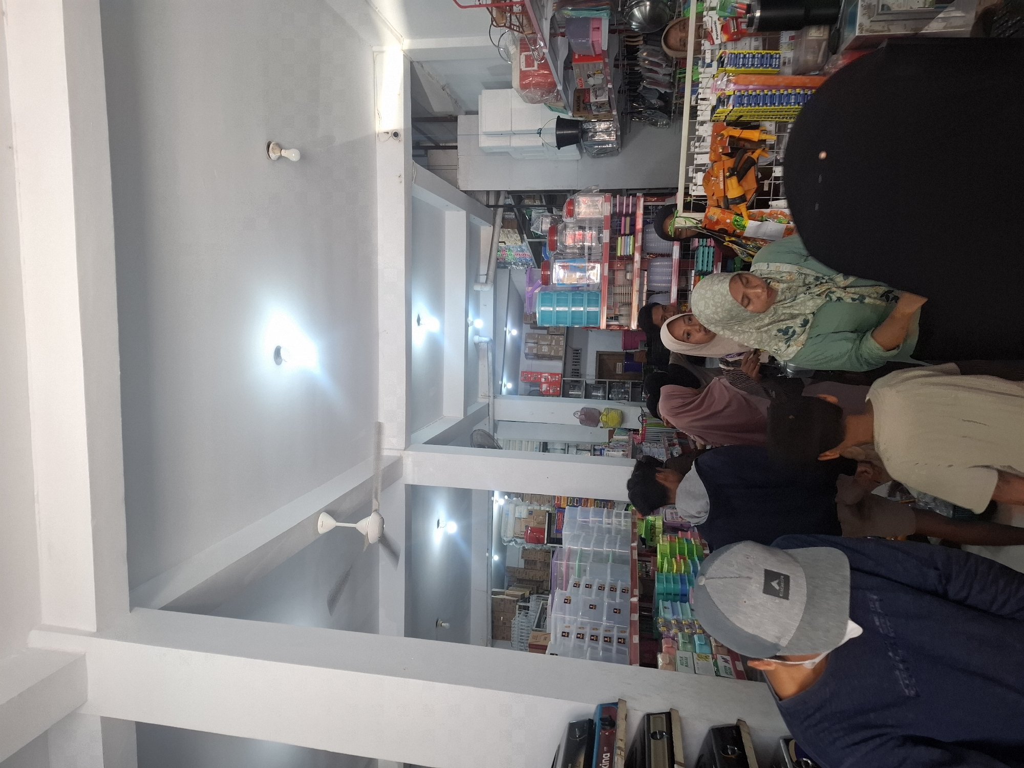 The image depicts the interior of a store, possibly a housewares or general goods store, in Sumberwudi, Indonesia. The store is filled with various products displayed on shelves and racks. In the foreground, a person wearing a blue long-sleeved shirt and a grey baseball cap is facing away from the camera. To their right, a young person in a light-colored t-shirt is also facing away. Further into the store, several other individuals, primarily women wearing headscarves, are visible browsing the merchandise. One woman in a green headscarf and green top is looking down at something in front of her. The shelves are stocked with items like plastic containers, kitchenware, and boxes. The lighting suggests it is daytime, with ceiling lights illuminating the space. The architecture includes white pillars and a white ceiling with a ceiling fan. The overall impression is a busy retail environment.