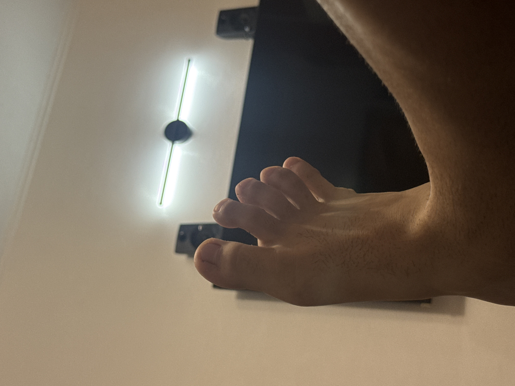 The image captures a close-up, low-angle perspective of a bare right foot and lower shin of an individual, positioned in an elevated and relaxed posture. The foot exhibits natural skin tone with visible fine hair on the shin and toes. The toes are slightly curled upwards. In the immediate background, a broad, dark horizontal surface is visible, likely a television screen or an entertainment unit. Situated on this dark surface, two dark, rectangular audio speakers are symmetrically placed, one to the far left and one to the far right. The upper background features a plain, light beige wall, against which a linear light fixture is mounted. This fixture consists of a dark circular central mount from which two horizontal, illuminated bar-like elements extend outwards, emitting a bright white light. The scene depicts an indoor environment, potentially a living room or bedroom, located in the city of Varna, Bulgaria.