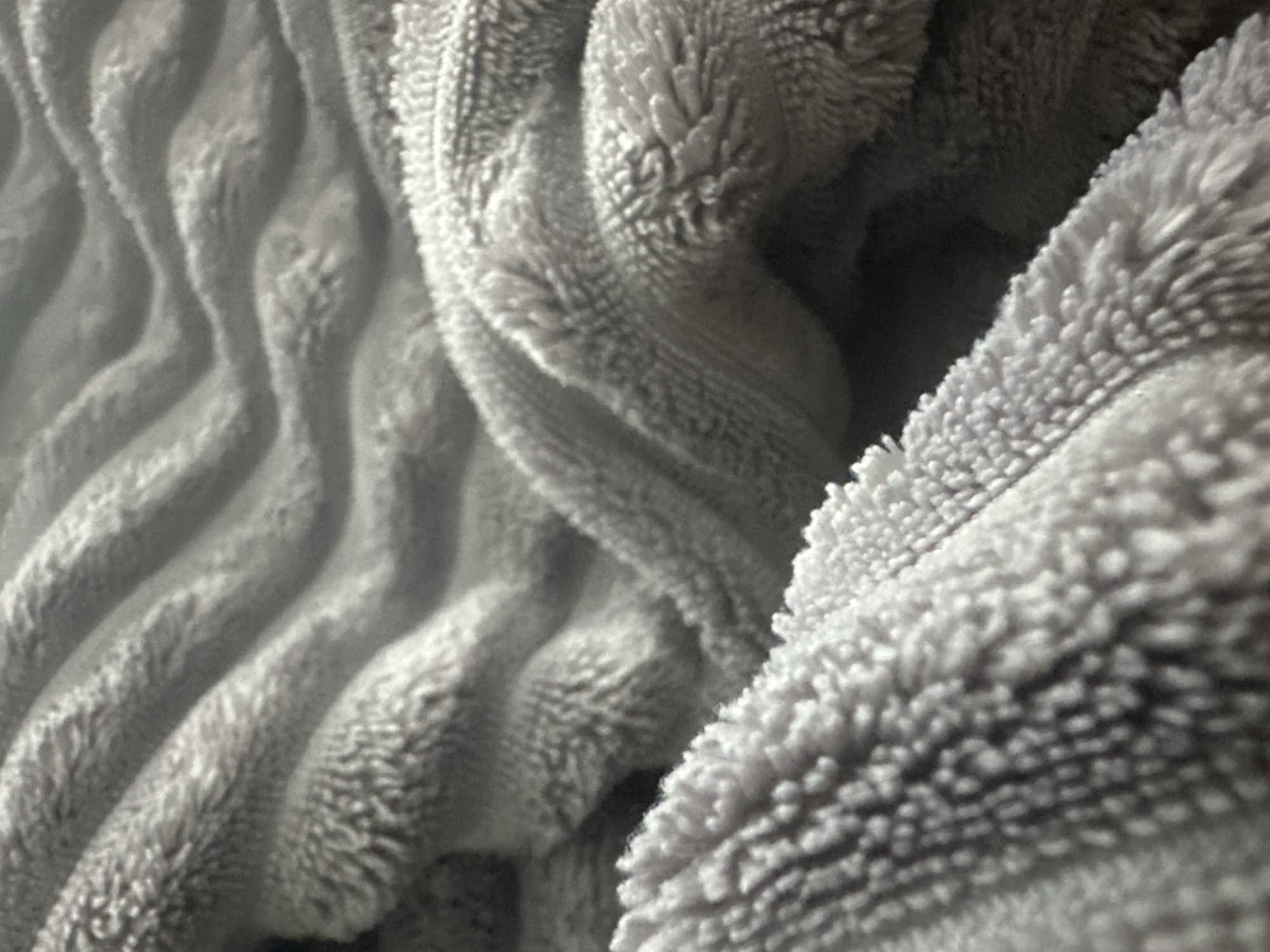The image is a close-up, abstract view of a textured, grey, plush fabric. The fabric displays a repeating ribbed pattern, creating a series of undulating waves or ridges. The texture is soft and fuzzy, with visible fibers that catch the light, giving it a slightly luminous appearance in some areas. The lighting creates shadows within the valleys of the ribs and highlights on the crests, emphasizing the depth and softness of the material. There are no discernible objects, people, actions, or interactions visible. The scene is entirely focused on the tactile and visual qualities of the fabric itself. There is no information within the image to suggest its location in Praskoveyevka, Russia.