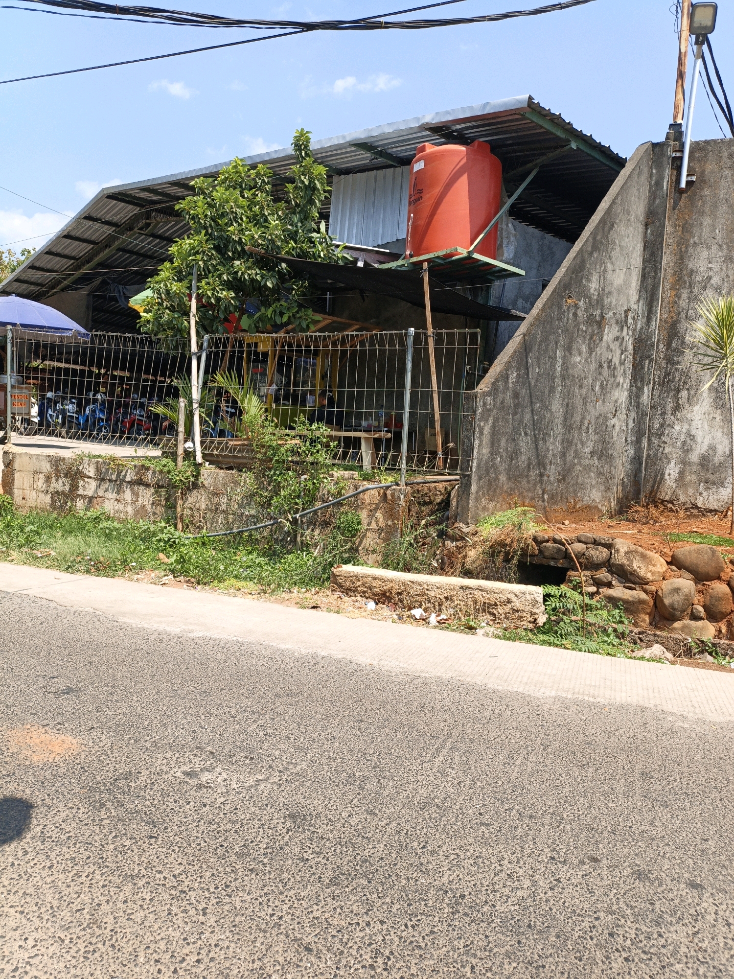 An outdoor, eye-level, medium shot captures a street-side view in Kuwasen, Indonesia, on a clear, sunny day. In the foreground, a paved road transitions to a concrete sidewalk bordering a slightly overgrown embankment. Beyond the embankment, a chain-link fence encloses an area with a covered structure. This structure, supported by metal poles, has a corrugated metal roof. An orange water tank sits on a metal platform atop the covered area, to the right of a large green tree. Behind the fence, several motorcycles are parked under the shelter. To the far right of the image, a tall, rough concrete wall rises, adorned with a streetlamp on a rusty pole. The wall casts a shadow onto the embankment, which is dotted with rocks and vegetation. The sky above is bright blue with a few scattered white clouds.