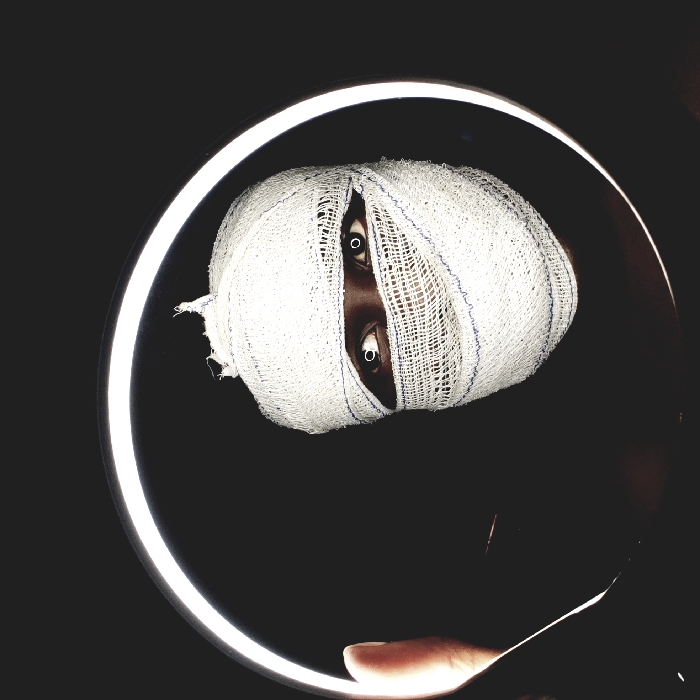 A person's head is wrapped in bandages, leaving only the eyes visible. The eyes are wide open and appear bright, possibly due to a light source reflecting in them. The head is framed within a circular light against a dark background. A hand partially enters the frame at the bottom, suggesting the subject is being held or positioned within the light. The overall effect is striking and potentially unsettling.