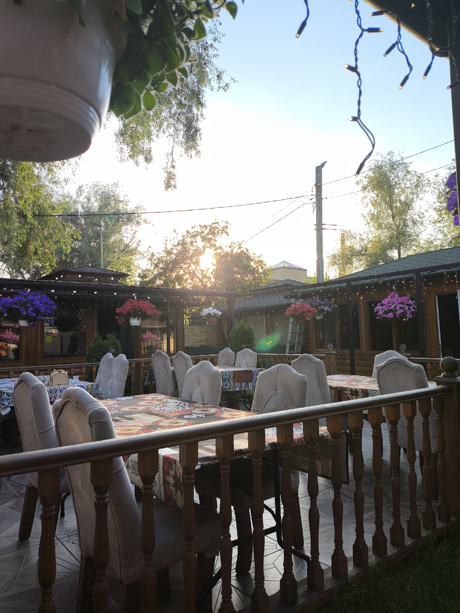 The image depicts an outdoor dining patio at an establishment located in the city of Unknown, Unknown, captured during what appears to be late afternoon or early evening due to the low sun. The scene is devoid of people.
In the foreground, a dark wooden railing with turned balusters spans the lower portion of the frame, separating a small patch of green grass on the immediate right from the patio area. Behind this railing, multiple rectangular dining tables are arranged, each covered with distinct, vibrantly patterned tablecloths featuring geometric and possibly ethnic designs in hues of red, orange, blue, and white. Beige or light grey upholstered chairs with high backs and wooden legs are positioned around the tables. The patio floor is tiled with a geometric pattern, possibly hexagonal or octagonal.
The middle ground is dominated by a single-story wooden building or a series of interconnected wooden structures forming the main part of the establishment. These structures have dark-stained wooden paneling, large windows, and dark-colored roofs. Strings of small, warm-white decorative lights are strung along the eaves and what appears to be a pergola structure overhead, suggesting illumination for evening use. Several large hanging baskets filled with blooming flowers in shades of deep purple, red, and pink are suspended from the overhead structure. Small potted evergreen shrubs are placed on the patio near the building.
In the background, a bright, hazy sky is visible, with the setting sun largely obscured by the dense canopy of mature deciduous trees, creating a strong backlighting effect and a lens flare near the center-right. A utility pole with overhead wires is visible against the sky on the left side, behind the patio structures. The very top-left corner features a partial view of a light-colored hanging planter with trailing green foliage. The overall environment is green and lush, with trees encircling the outdoor dining area.