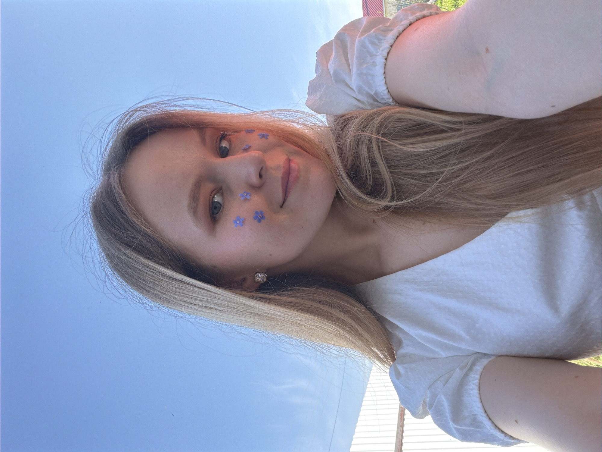 A young woman with fair skin and blonde hair is facing the camera. Several small blue flowers are adhered to her left cheek and near her left eyebrow. She is wearing a white blouse with short, puffed sleeves. A square earring is visible on her left ear. The background consists of a clear blue sky and a portion of a building or structure made of light-colored, possibly white, materials. The location is possibly Кадряково, Russia.