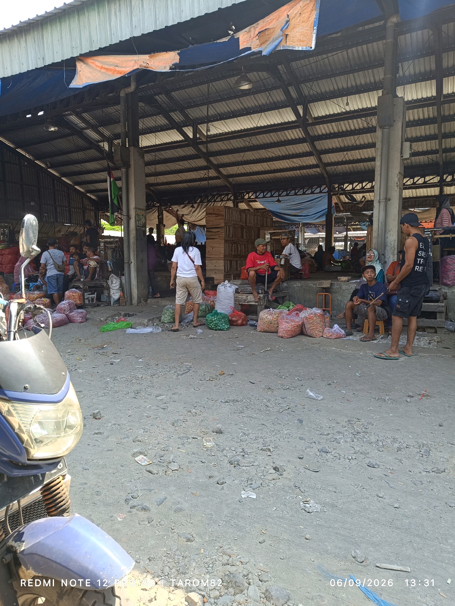 The image captures a bustling outdoor market scene, likely in Pare, Indonesia, given the date and time stamp suggesting a local context. The market is under a large, open-sided structure with a corrugated metal roof, partially covered by blue and orange tarps. The ground is unpaved and appears to be a mixture of dirt and gravel, with some scattered debris.
Several individuals are present, engaged in various activities. In the foreground, a blue motorcycle is parked, partially obscuring the view of the market floor. Beyond the motorcycle, numerous sacks and piles of produce, predominantly red chilis and green vegetables, are displayed for sale.
In the middle ground, a woman in a white shirt and khaki shorts stands facing away from the camera, possibly interacting with a vendor or customer. To her left, a group of people are seated on low benches, some appearing to be vendors or shoppers taking a break. Further to the left, more people are visible standing and milling about amongst the goods.
On the right side of the image, a man in a black tank top and shorts stands conversing with a seated individual. Other figures are scattered throughout the market, adding to the lively atmosphere. The overall impression is of a typical Indonesian wet market, characterized by its open-air setting, abundance of fresh produce, and the presence of a diverse community. The date and time stamp, "06/09/2026 13:31," along with the text "REDMI NOTE 12 PRO 5G TAROM82," indicates the photograph was taken on September 6, 2026, at 1:31 PM, using a Redmi Note 12 Pro 5G phone.