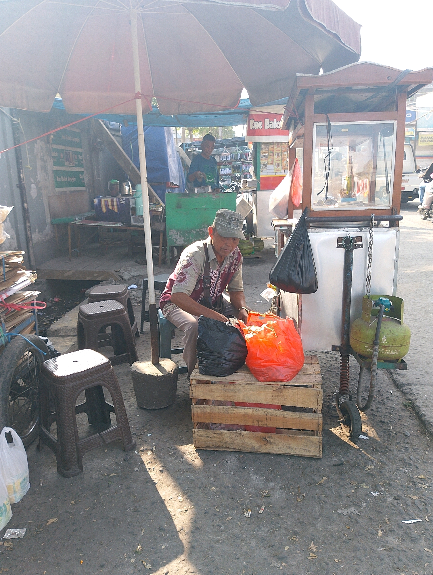 The image depicts an outdoor scene in Lembang, Indonesia, likely a street market or roadside stall area. The primary subject is a middle-aged man, wearing a cap and a patterned shirt, sitting on a stool next to a wooden crate. He is handling two large plastic bags, one black and one orange.
Behind him, a vendor cart with a glass display case and a prominent umbrella is visible. The cart appears to be selling snacks or food items, with signage in Indonesian that includes "Kue Balok." A green propane gas tank is attached to the side of the cart, suggesting cooking is involved.
The environment is somewhat cluttered, with various objects scattered around. These include other stools, a bicycle wheel, stacks of cardboard or paper, and debris on the ground, indicating a busy, possibly less organized, commercial area. The lighting suggests it is daytime, with sunlight casting shadows. Another person, a man in a dark shirt, is partially visible in the background, interacting with the vendor cart. The overall impression is of a typical street scene in a busy Indonesian town.