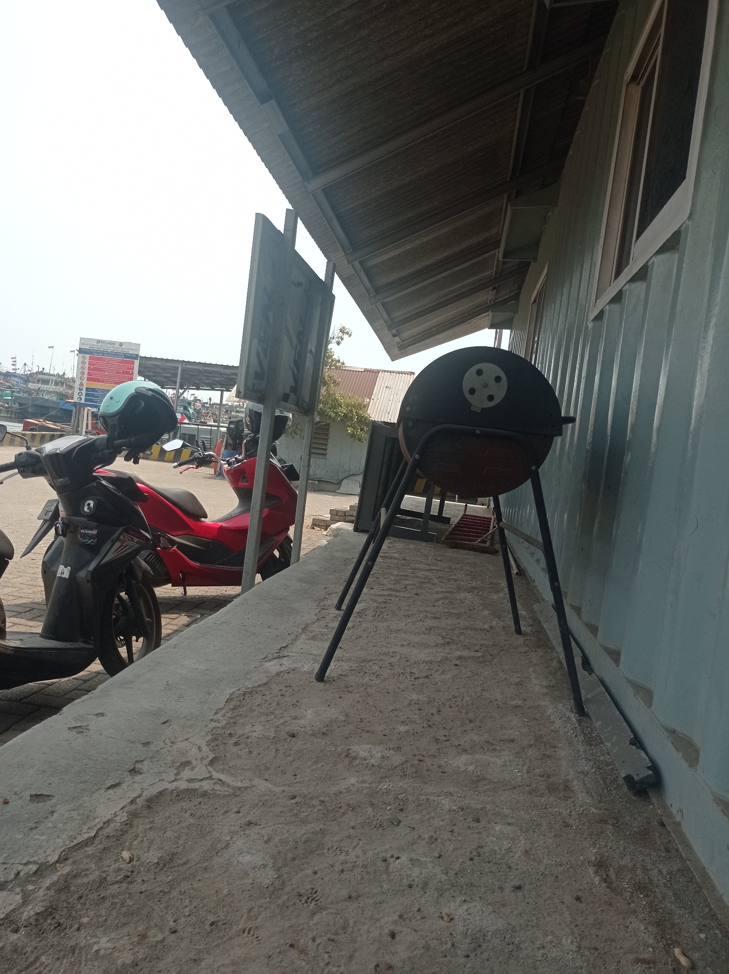 The image depicts an outdoor scene in Tegal, Indonesia, likely near a harbor, evidenced by the distant boats and a sign that appears to be for a port authority. In the foreground, a black and a red scooter are parked on a concrete surface. A light blue helmet rests on the handlebars of the black scooter. To the right, a black barbecue grill with three legs stands on the ground. Behind the scooters and grill is a building with a corrugated metal roof and light blue corrugated metal siding. The ground is a mix of concrete and sandy dirt. The sky is bright and overcast.