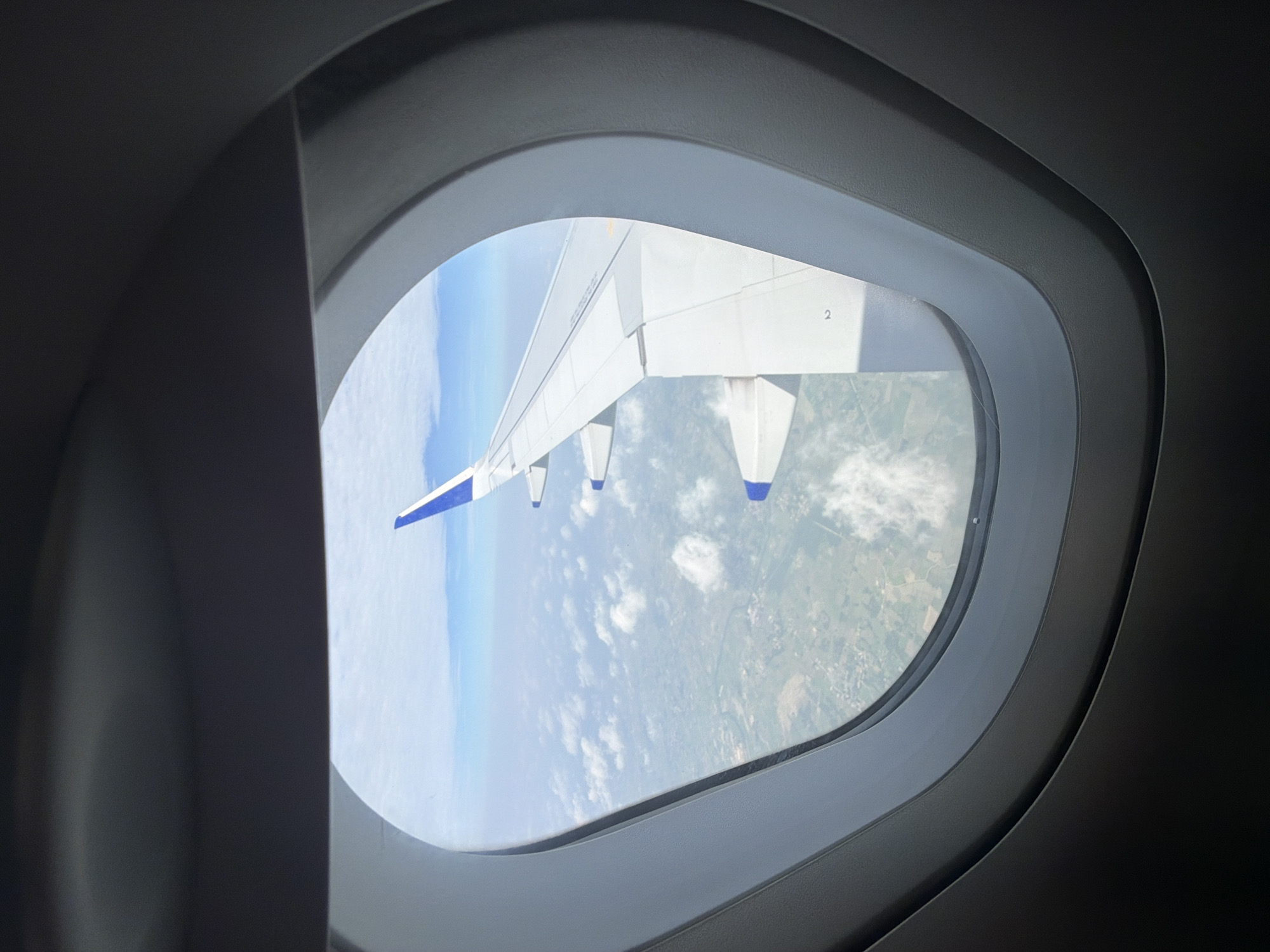 A view from an airplane window shows the wing of the aircraft extending to the right. Below the wing, a patchwork of green fields and scattered buildings is visible, indicative of the landscape surrounding Mohan, India. The sky is a bright blue, dotted with scattered white clouds. The interior of the airplane cabin is dark and frames the window view.