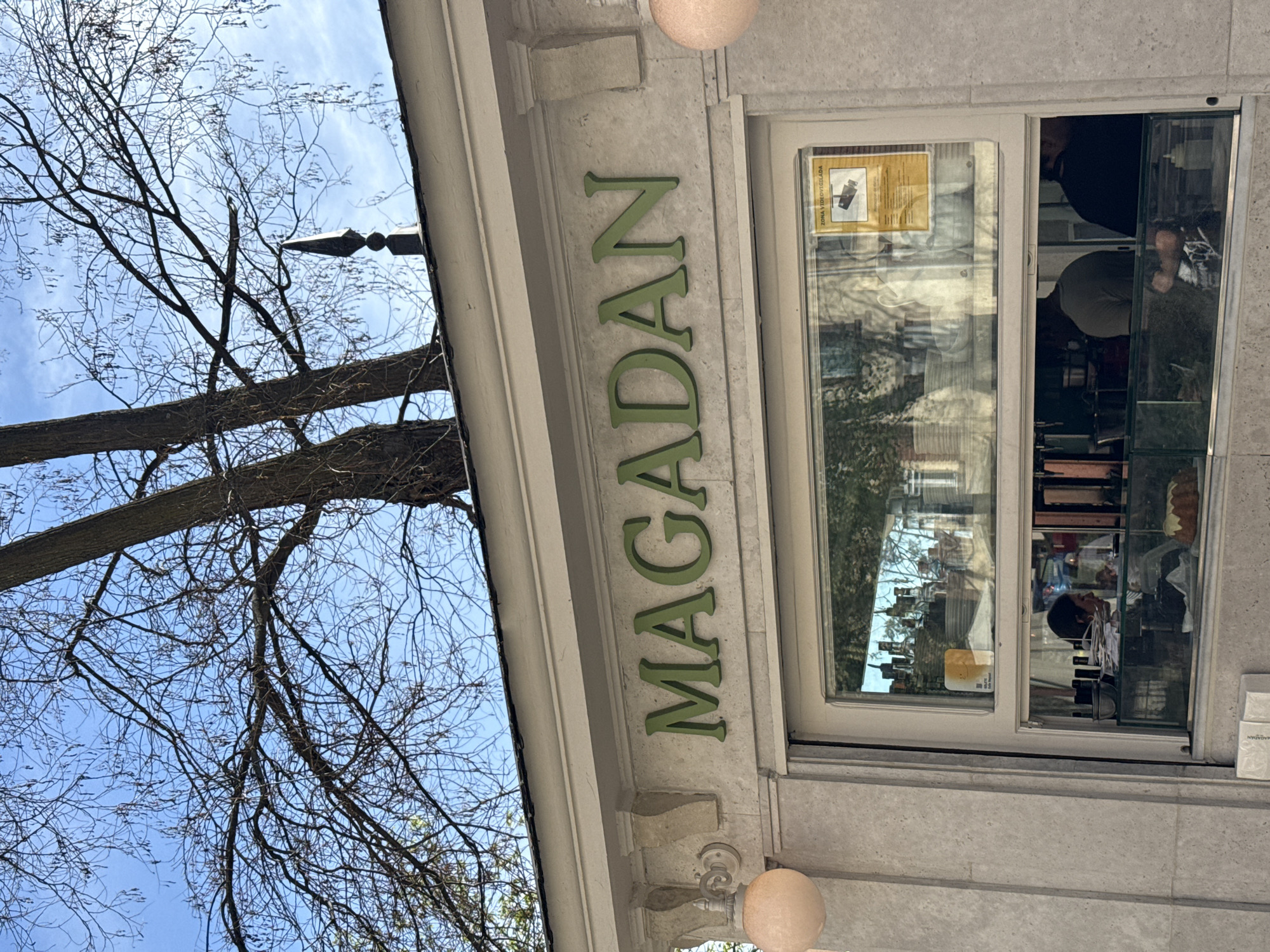 The image shows the storefront of "Magadan," a business located in Madrid, Spain. The sign "MAGADAN" is displayed in green letters above the storefront. A window is visible, reflecting the surrounding environment, including trees. Inside the storefront, people are present, and various items are on display. A tree with bare branches is also visible in the background against a blue sky.