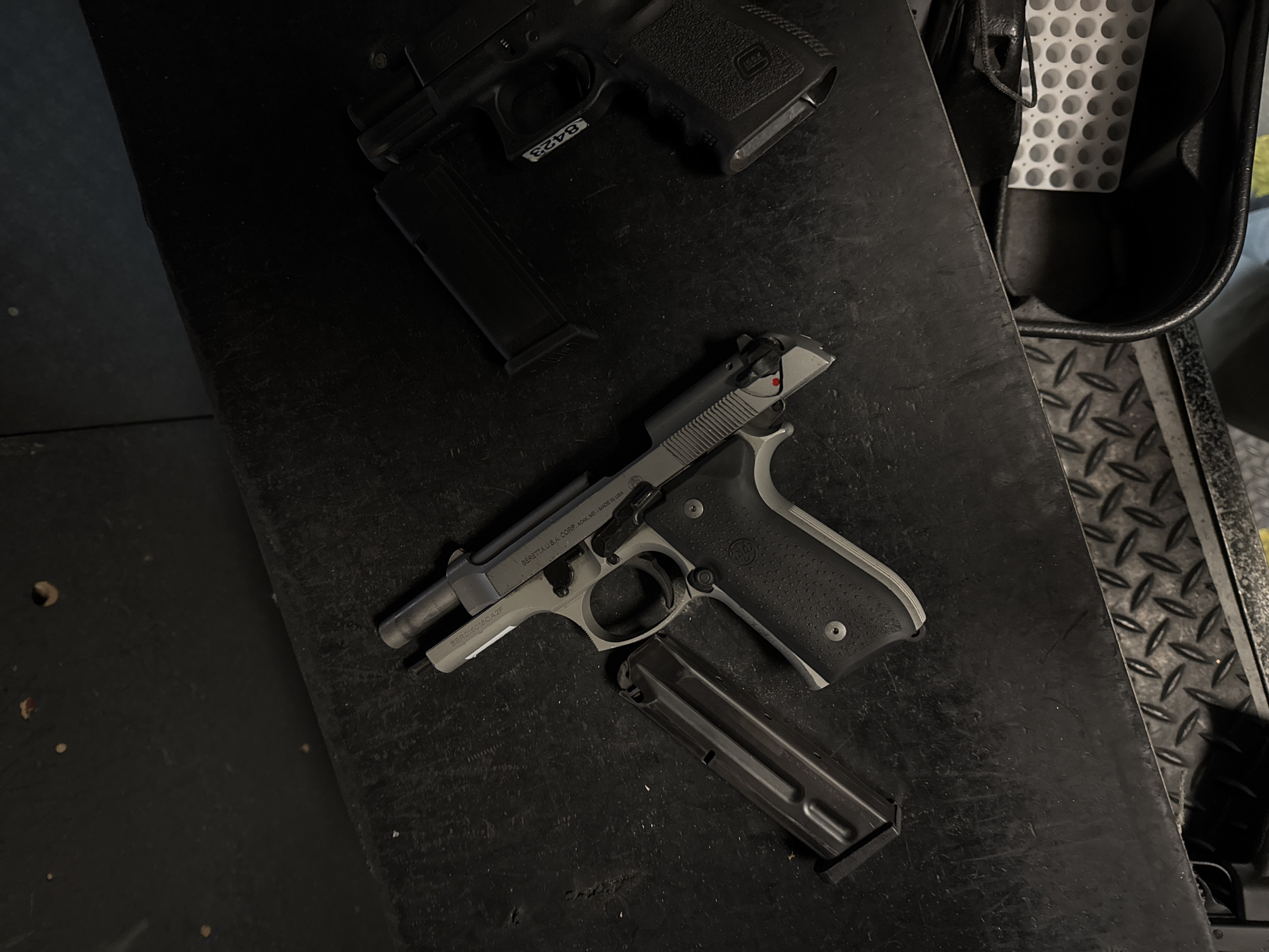The image shows two firearms and magazines lying on a dark, possibly metallic, surface. The upper firearm is a black Glock pistol with a magazine inserted, and a partially visible label with the number "8423". The lower firearm is a Beretta pistol, silver and black, with a magazine lying partially inserted beneath it. The words "BERETTA U.S.A. CORP. ACKK, MO MADE IN USA" and "BER0150180A2F" are visible on the slide of the Beretta. Next to the firearms, a container with a grid pattern is visible on the right. The scene is not in the city of Los Angeles and the image does not provide any information about people, actions or interactions.