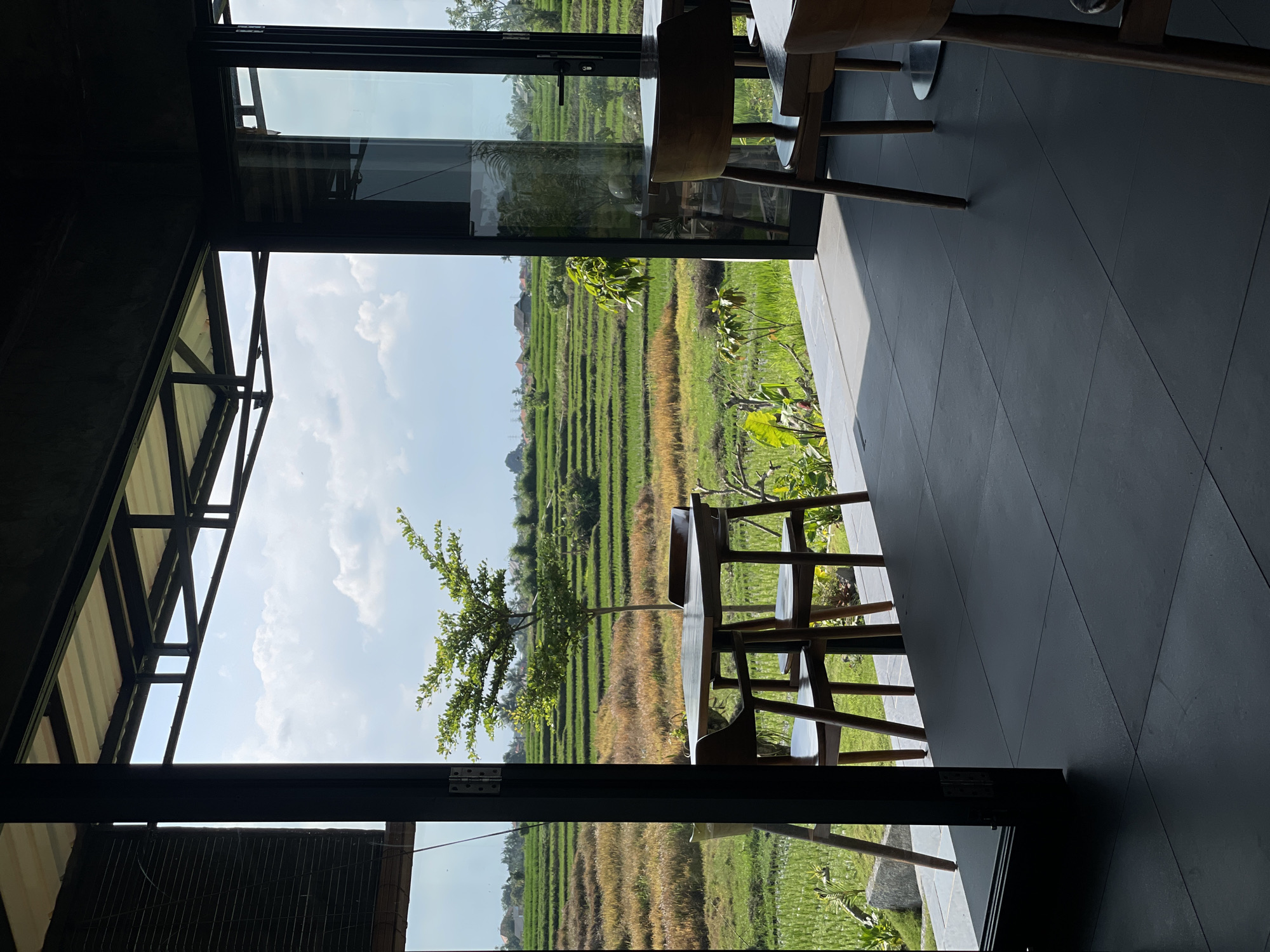 A view from inside a modern cafe or restaurant shows a table with two chairs placed in front of a large open window overlooking terraced rice fields in Kediri, Indonesia. The interior features dark tiled flooring and dark-framed glass doors. Outside, the lush green and golden-brown rice paddies are illuminated by bright sunlight, with a clear blue sky and scattered white clouds above. The scene conveys a tranquil atmosphere, likely during daylight hours.