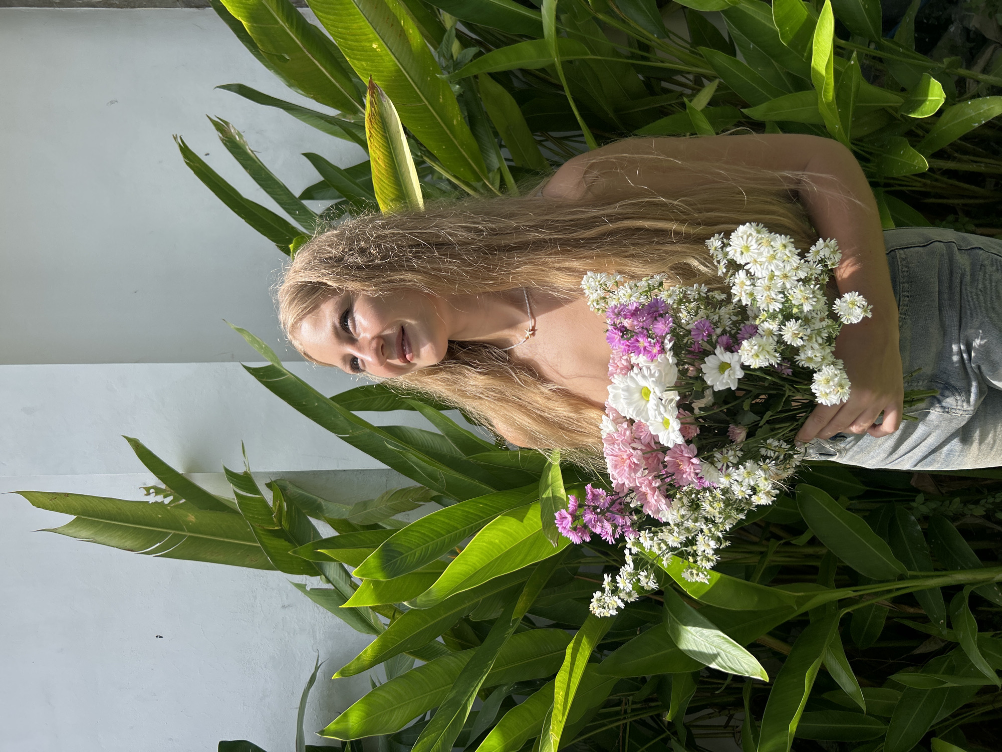 The image features a young woman with long, flowing blonde hair, captured outdoors against a backdrop of a white wall and lush green tropical foliage. She is smiling softly at the viewer, her head tilted slightly. She wears a light-colored top, possibly strapless, and light-wash denim jeans or shorts, with a delicate necklace adorned with a small charm visible around her neck.
In her arms, she holds a generous bouquet of wildflowers composed of white, various shades of pink, and purple blossoms, including small daisy-like flowers and larger white chrysanthemums. The flowers appear fresh and vibrantly colored.
The setting is brightly illuminated by natural daylight, creating clear highlights on the leaves and casting distinct shadows on the white wall behind her. This suggests a sunny day, likely during morning or late afternoon. The abundant, large-leafed tropical plants strongly indicate a warm, exotic climate, aligning with the specified location of Kuta Selatan, Indonesia. The woman's expression conveys contentment and happiness. There is no visible text within the image.