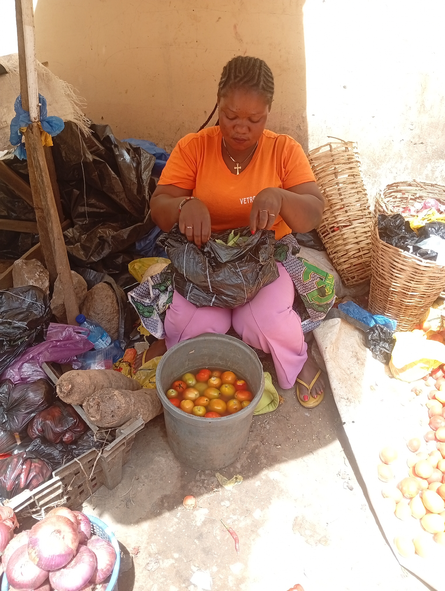 The image shows a woman in an orange t-shirt sitting in a market stall surrounded by produce. She is holding a black plastic bag and looking down at it. In front of her is a bucket of tomatoes soaking in water. To her left are yams, onions, and more plastic bags. Behind her are baskets, some filled with black plastic bags. The market stall appears to be constructed with wooden supports and covered with a tarp. The location appears to be Unknown, Unknown.
