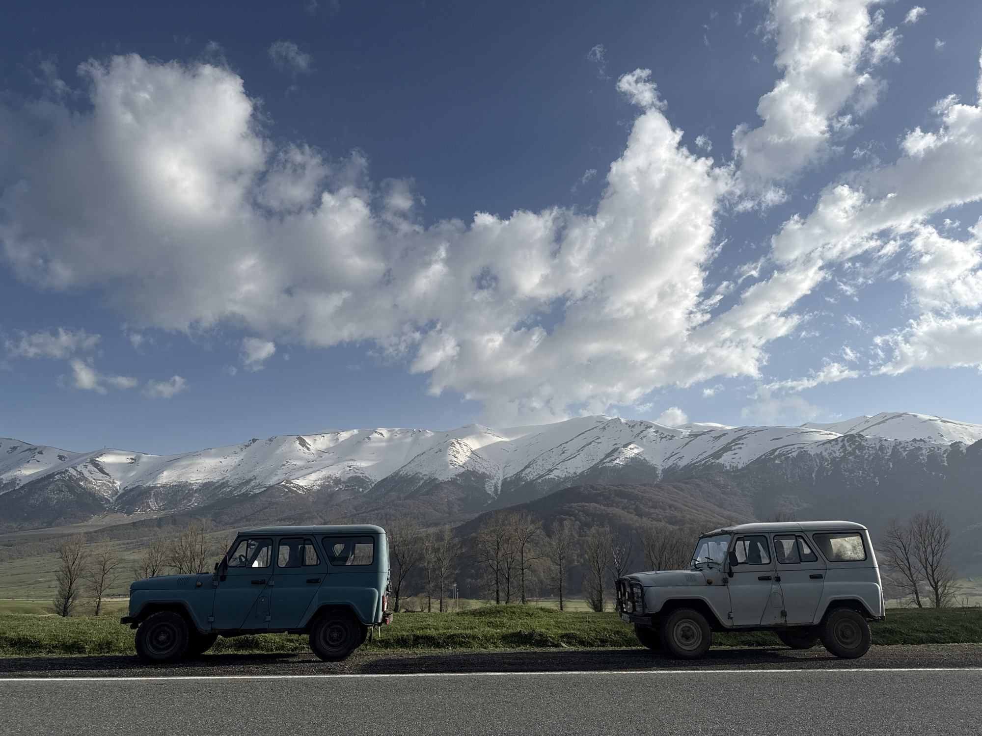 Two vintage UAZ-469 vehicles are parked on the side of a paved road. The vehicle on the left is blue-green, while the one on the right is gray. A grassy shoulder lies between the road and the base of snow-capped mountains in the background. Sparse, bare trees are scattered across the landscape. The sky is blue with scattered white clouds. This image was taken in or near Margahovit, Armenia.