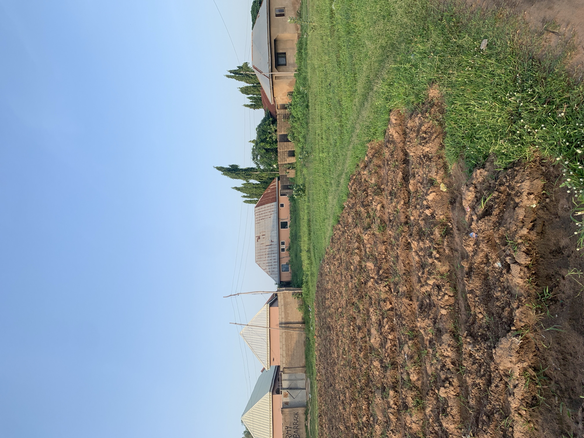 The image depicts a view of a residential area in Jalingo, Nigeria, under a clear blue sky. In the foreground, a patch of tilled earth shows rows of soil, indicating it is a garden or field. To the right, lush green grass with scattered white wildflowers covers the ground.
In the middle ground, a low brick wall separates the cultivated area from the residential properties. Beyond the wall, several houses with distinctive A-frame roofs are visible. One house on the far left has "ARMY BARRACK" spray-painted on its exterior wall. To the right of the houses, tall, slender trees stand in a line, forming a natural boundary.
The houses are of simple construction, with one appearing to be made of mud or adobe with a corrugated metal roof. Windows are visible on some of the structures. Electrical poles and wires are present, suggesting the presence of utilities. The overall scene conveys a sense of a developing or established residential neighborhood in a semi-urban or rural setting.