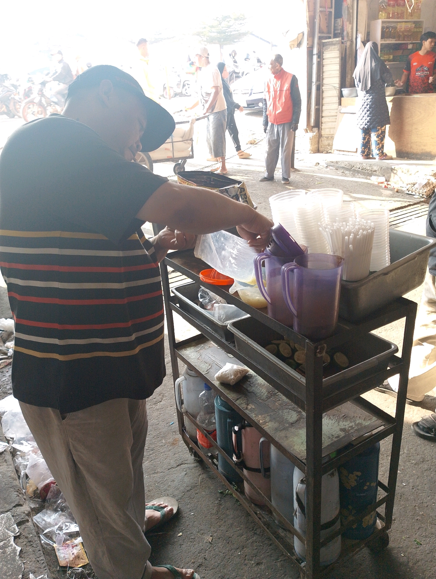 A street vendor in Lembang, Indonesia, is preparing drinks. The scene appears to be an outdoor market or roadside stall. The vendor, wearing a horizontally striped t-shirt and light-colored pants, is reaching into a metal cart filled with containers and cups. On the cart's shelves are stacks of clear plastic cups, a tray of lime slices, and two purple pitchers. Below, the cart holds several large cylindrical containers, possibly for water or other beverages. In the background, other people are visible, some walking and one standing near a shop counter. Motorcycles are parked nearby, suggesting a busy area. The ground is paved with concrete and littered with plastic bags and debris. Sunlight casts strong shadows across the scene.