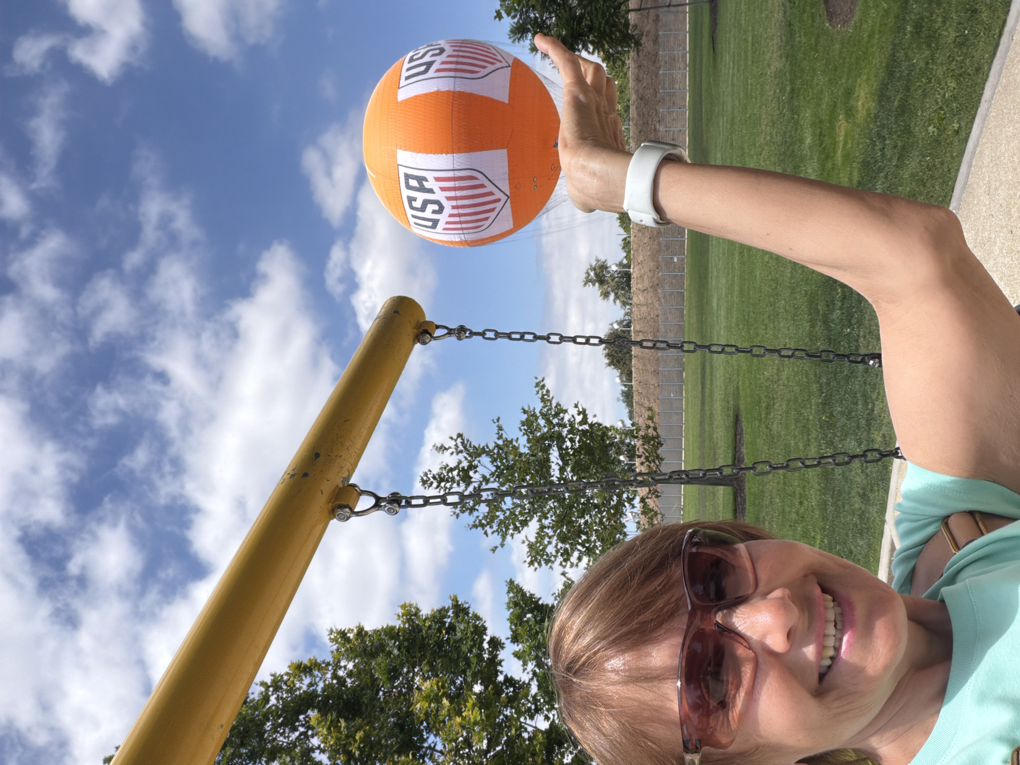 A woman smiles at the camera while holding an orange and white inflatable ball with "USA" logos on it. She is wearing sunglasses and a teal t-shirt. She has a white fitness tracker on her left wrist. The ball is suspended from a yellow swing set beam by a chain. The background is a park with green grass and trees under a blue sky with white clouds. The location is likely a park in Irvine, California, given the presence of a playground.
