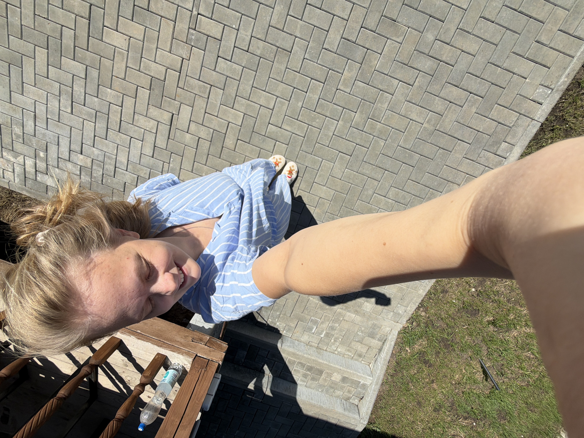 The image, taken from a high angle, shows a woman in a blue and white striped shirt standing on a brick-paved area with a grassy section visible on the right. She is wearing patterned slip-on sandals. Part of a wooden railing with a water bottle on it is visible in the lower left corner. The photograph appears to be a selfie, as the arm holding the camera is in the foreground. The location is likely outdoors and in a residential area, but specific details to confirm it is Kamennoye, Russia, are not present in the image.