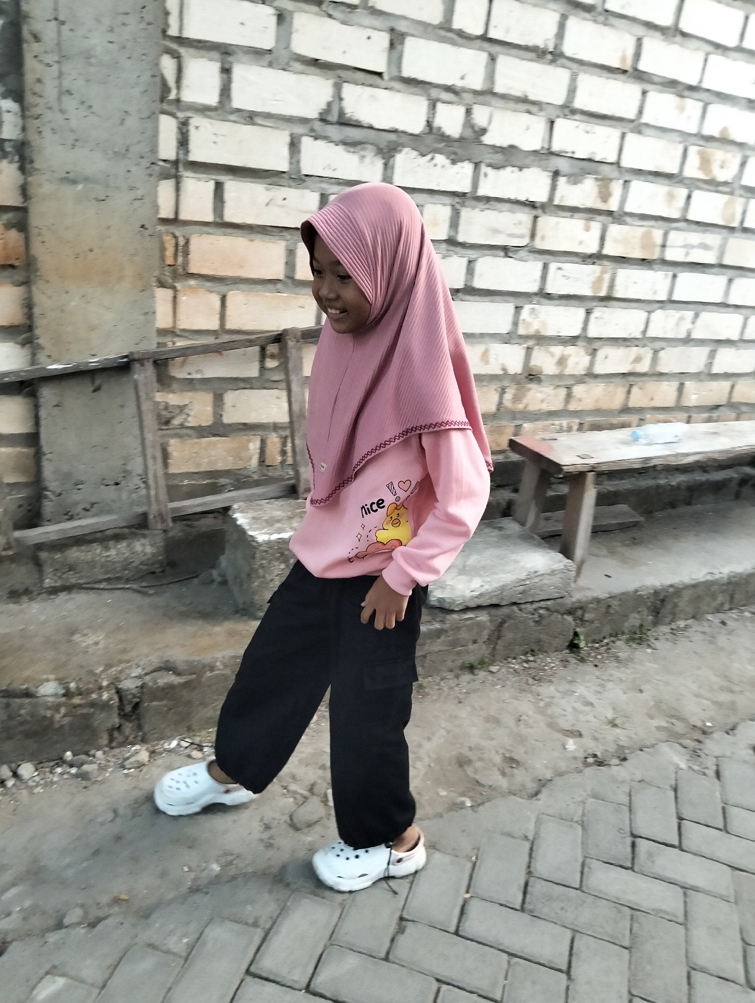 A young girl wearing a pink hijab and pink sweatshirt, black pants, and white slip-on shoes is standing on a brick-paved sidewalk. She is smiling and looking to her right. In the background, there is a brick wall, a wooden bench, and some construction debris. The scene appears to be outdoors in a semi-urban environment, possibly in Paciran, Indonesia.