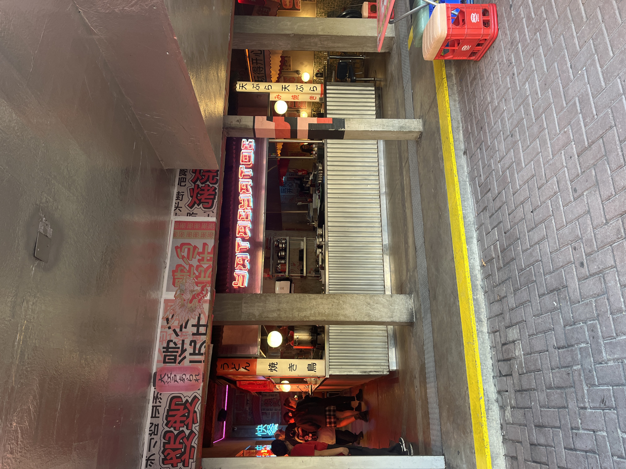 The image shows a narrow alleyway with Japanese restaurants and signage on both sides. People are gathered at the end of the alleyway. The restaurants have corrugated metal shutters. A red plastic crate is on the right side of the alleyway, next to a yellow curb. The alleyway is paved with bricks. Many of the signs are in Japanese.