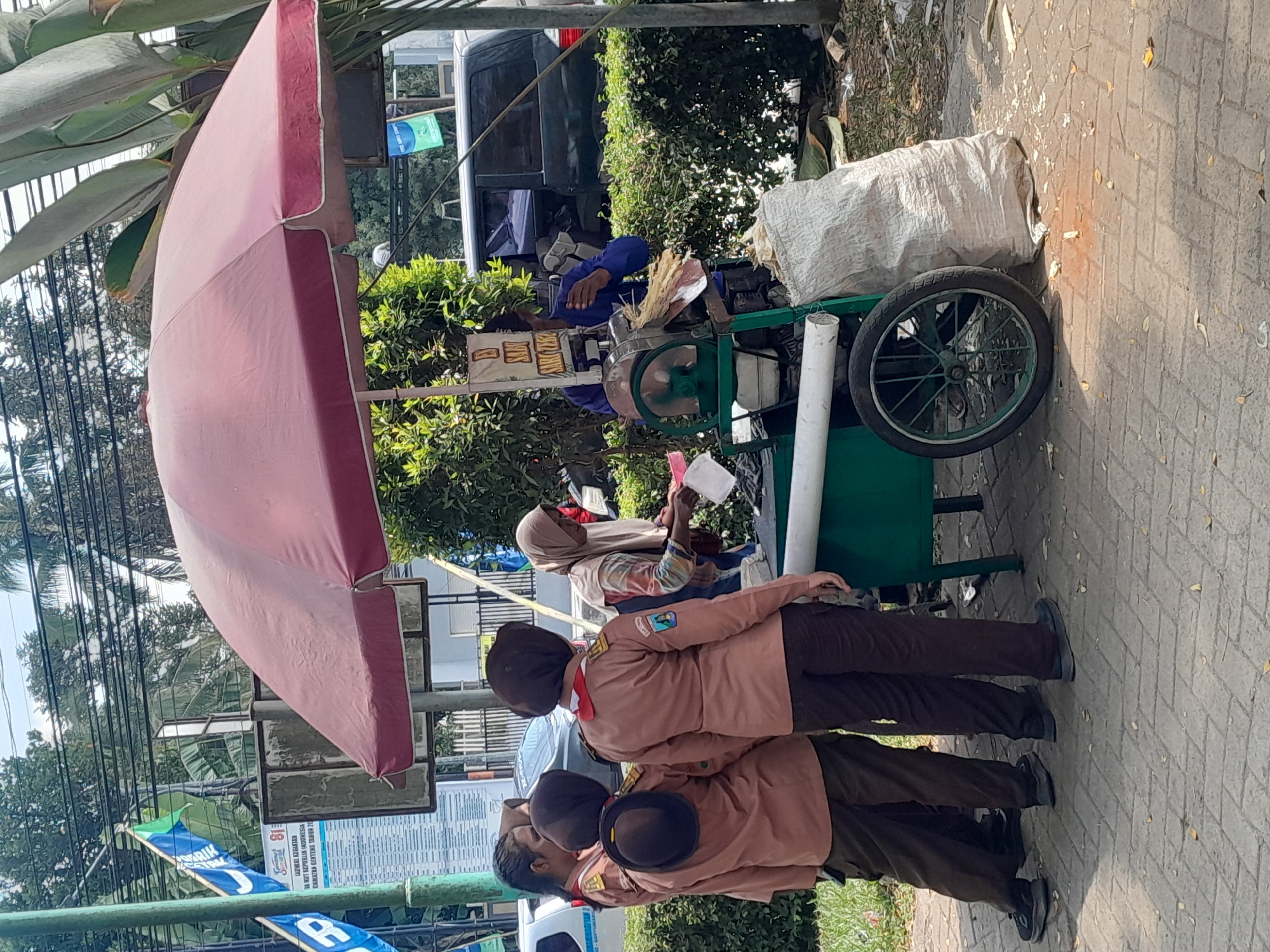 In Genteng Kulon, Indonesia, an outdoor scene depicts a street vendor operating a sugarcane juice machine under a large red umbrella. Three young females in brown scout uniforms stand to the left of the machine, facing away from the camera, appearing to be observing or interacting with the vendor. A female vendor, wearing a headscarf and a patterned shirt, is positioned behind the machine, actively engaged with it. The sugarcane juice machine is green, with a wheel on the left side and a large sack of what appears to be sugarcane pulp or waste to the right. In the background, a dark-colored SUV is parked, and various trees and foliage create a natural backdrop. Banners with Indonesian text are visible on poles to the left, suggesting a public or commercial area. The ground is paved with bricks, with scattered leaves and debris.