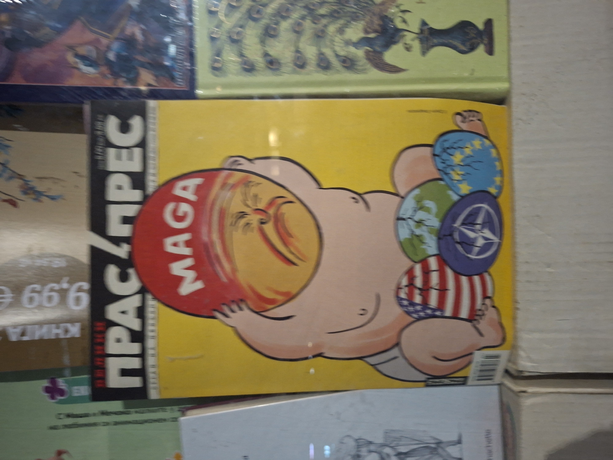 The image shows a satirical magazine cover with a caricature of Donald Trump as an obese baby. The baby has a red "MAGA" hat covering its head. In one hand, it is holding a globe, a European Union flag, a NATO logo, and the American flag. The magazine title, in Bulgarian, is printed along the side. The magazine is stacked with other books and magazines. The location is likely a bookstore or newsstand in Plovdiv, Bulgaria.