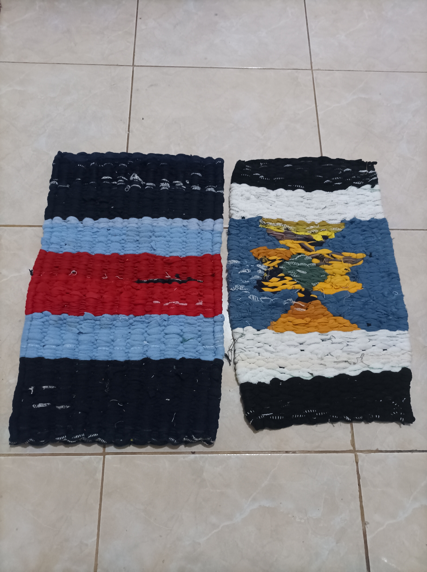 Two handmade woven rugs are displayed side-by-side on a tiled floor. The rug on the left features horizontal stripes of black, light blue, and red. The rug on the right depicts a colorful scene, possibly a landscape with a tree or abstract shapes, against a background of blue and white. The floor is composed of large, light-colored tiles with visible grout lines. The scene appears to be indoors, with no indication of an outdoor environment. The image does not provide any information to suggest a location in Nganjuk, Indonesia.