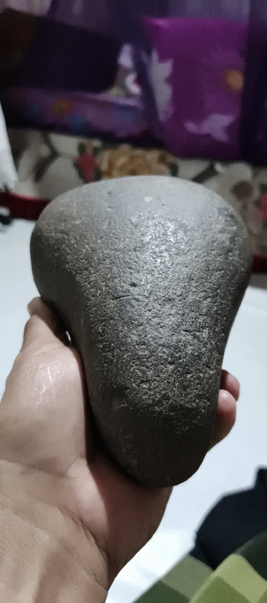 In Muaro Jambi, Indonesia, a hand is holding a dark gray, irregularly shaped stone that resembles a saddle or a smooth, flattened egg. The stone has a rough, textured surface with visible small pits and some faint lines or scratches. The hand holding the stone is light-skinned, with fingers curled around the object. In the background, there is a blurred area with purple fabric featuring white floral patterns, suggesting an indoor setting. The flooring is light-colored and reflective, indicating it might be tiles. The lighting appears to be from above, casting a subtle sheen on the stone and the hand.