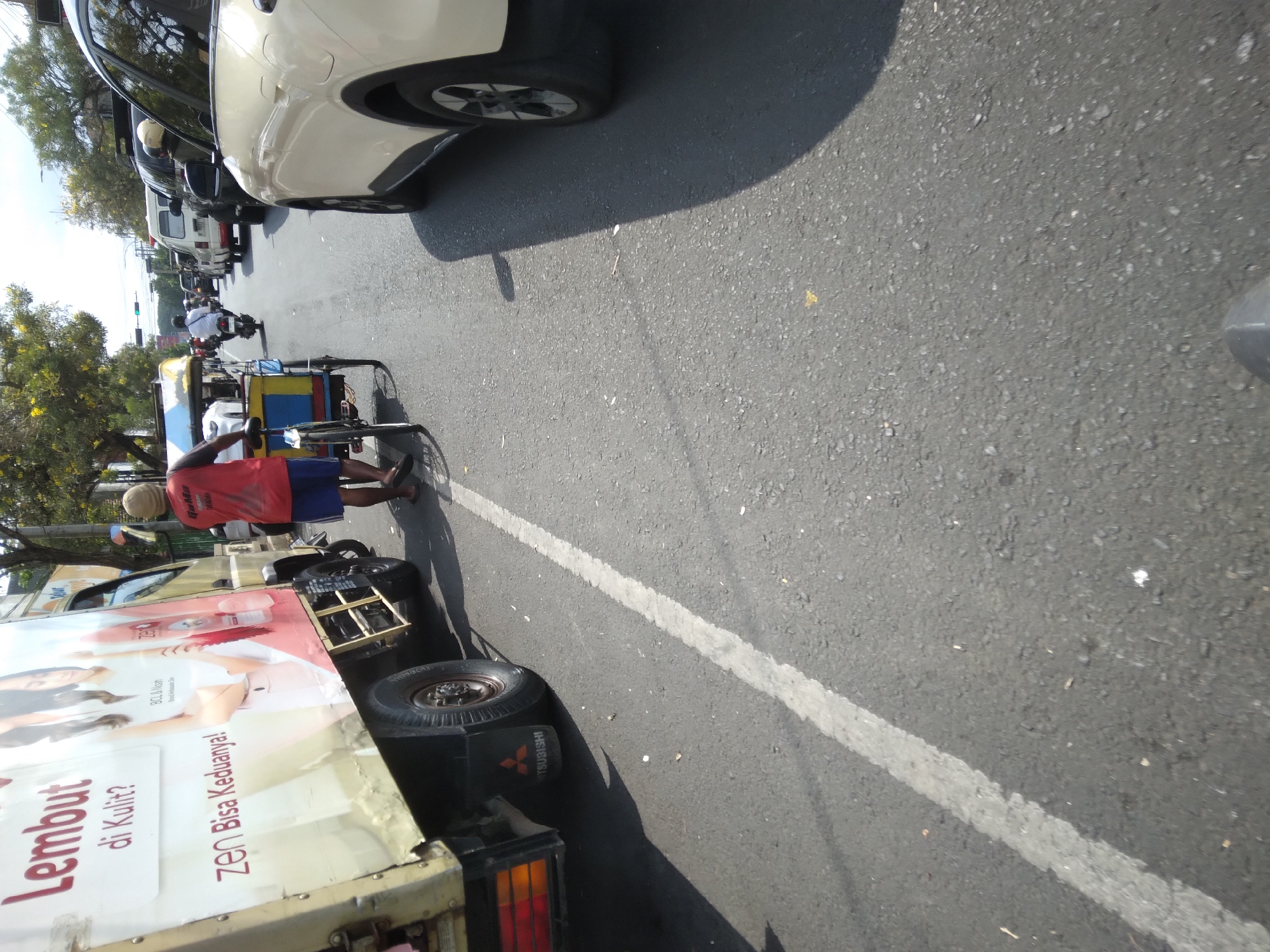 The image depicts a street scene in Purwokerto, Indonesia, characterized by moderate traffic. On the left, a yellow truck with large advertisements on its side is prominently featured. The advertisements are in Indonesian, with text such as "Lembut di Kulit?" (Soft on the Skin?) and "ZEN Bisa Keduannya!" (ZEN Can Do Both!). The truck has a Mitsubishi logo visible on its rear fender. To the right of the truck, a man wearing a red shirt and blue shorts is walking alongside a pedicab. The pedicab has a blue and yellow canopy. In the background, other vehicles, including cars, motorcycles, and a white van, are visible, suggesting a busy urban environment. Trees line the street, and the sky is bright, indicating daylight. The road surface is asphalt, with a white line marking a lane. The perspective of the image is low, as if taken from a moving vehicle or close to the ground. The overall impression is of a typical street scene in a Southeast Asian city, with a mix of commercial vehicles, public transport, and personal vehicles navigating the road.
