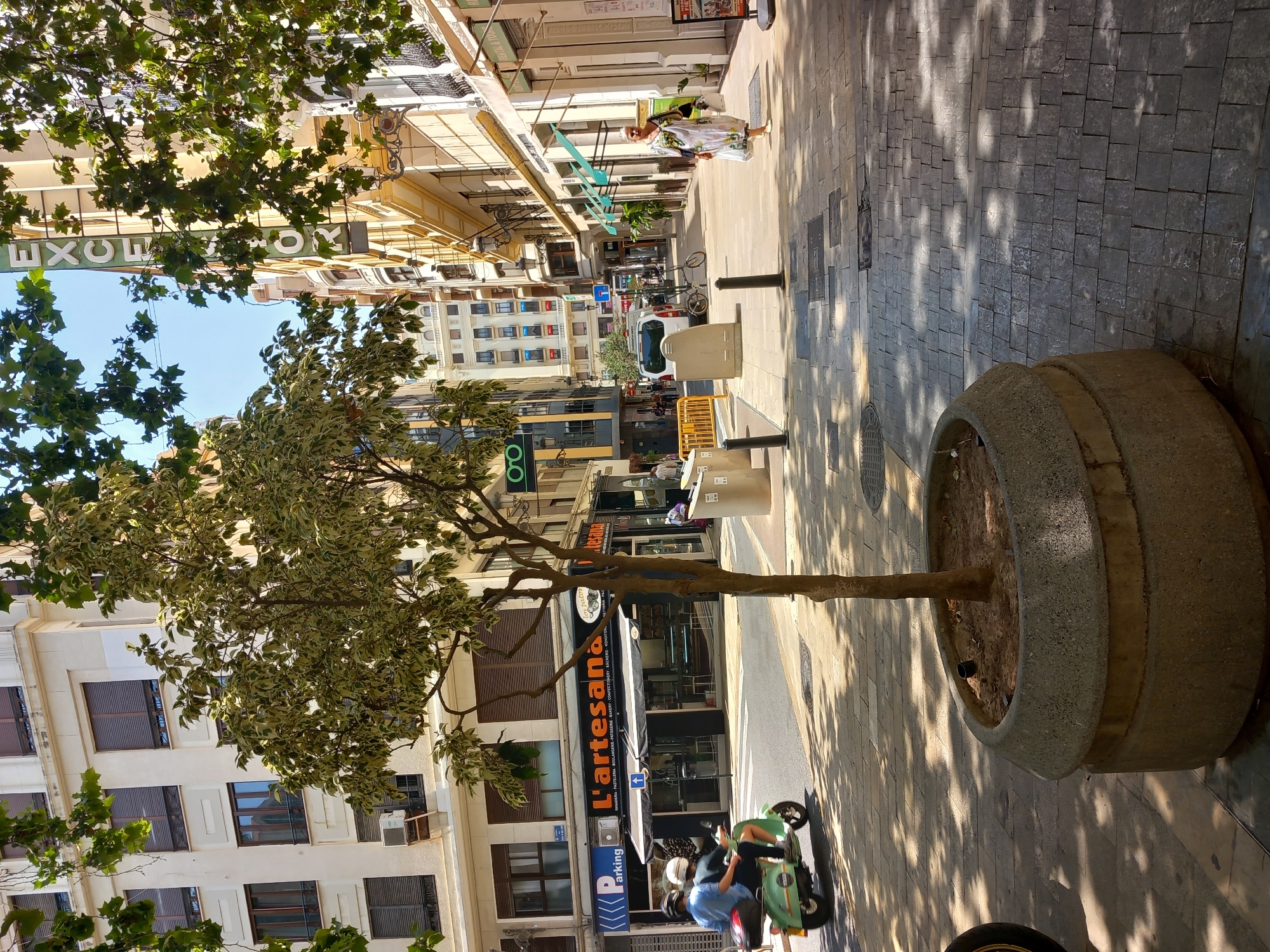 This is a street-level, eye-level, medium shot image of a pedestrianized street in Valencia, Spain, captured on a sunny day. In the foreground, a large, round, concrete planter contains a slender tree with variegated green and white leaves. The tree's shadow is cast across the textured, gray cobblestone sidewalk.
To the left, a light green scooter with two occupants wearing helmets is moving away from the camera on the asphalt street. Behind the scooter, a storefront with a black awning displays the name "L'artesana" and signage indicating it is a "PANADERIA, PASTELERIA, BOULANGERIE, PATISSERIE, BAKERY, CONFECTIONERY, BÄCKEREI, KONDITORI". Adjacent to it is a "Parking" sign.
The buildings lining the street are several stories high, with architectural styles common in European cities. On the right side of the street, a woman in a long, patterned dress walks away from the camera, carrying shopping bags. Further down the street, a white van is parked, and a bicycle leans against a barrier. A vertical sign with green lettering spelling "EXCELSIOR" is visible on a building further down the right side of the street, suggesting a hotel or business establishment. The sky is clear and blue. The overall scene depicts a typical urban street environment with shops, pedestrians, and vehicular traffic in Spain.