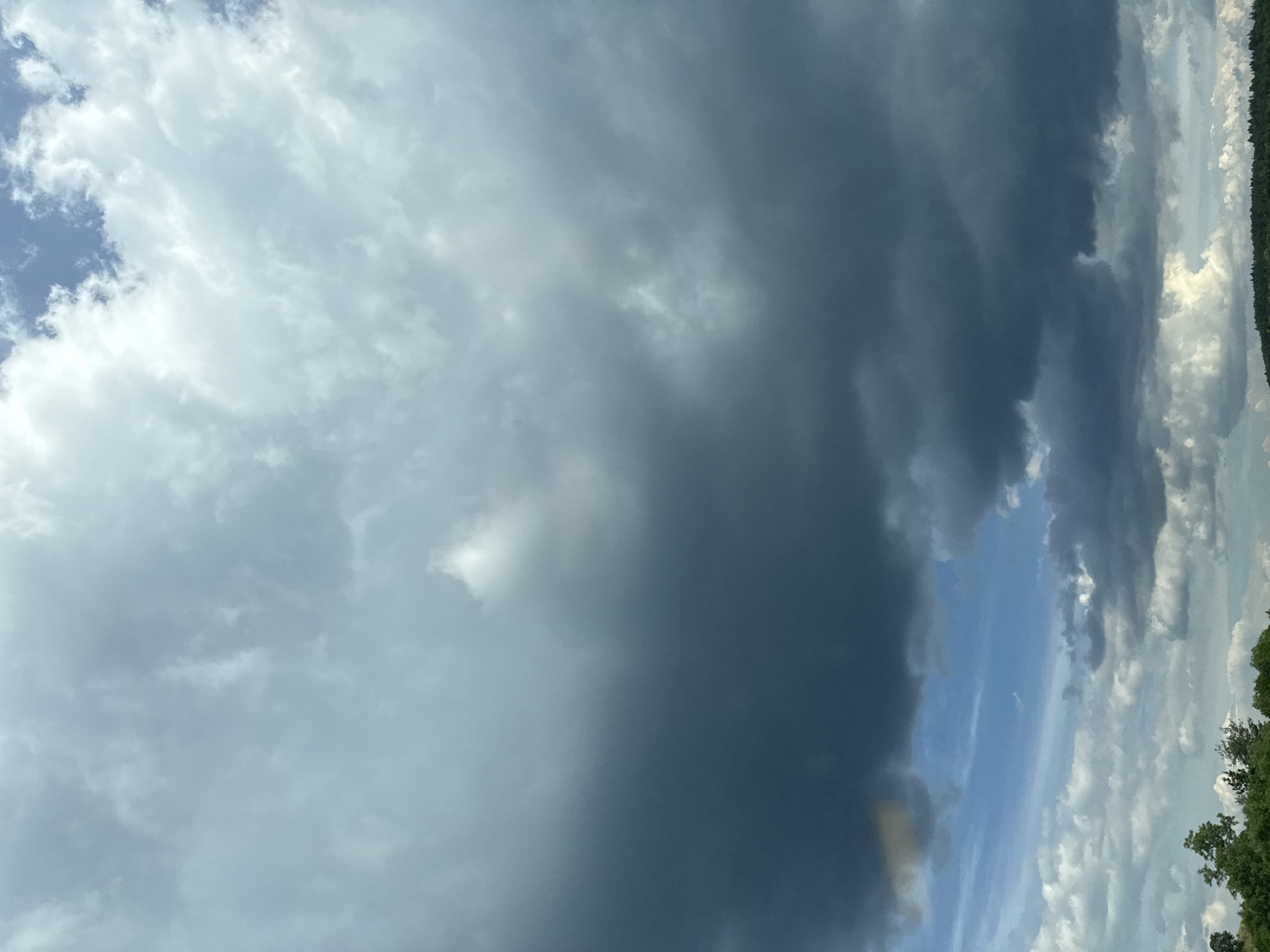 This is a photograph of a stormy sky over a landscape. The majority of the image is filled with dark, heavy clouds, suggesting an impending storm. There are breaks in the clouds revealing patches of blue sky. The bottom portion of the image shows a tree line with some greenery, hinting at land, possibly hills or a forest. There is no visible evidence of people or specific structures to indicate the location is the city of Rajana, Bulgaria. The lighting is dim and overcast due to the cloud cover.