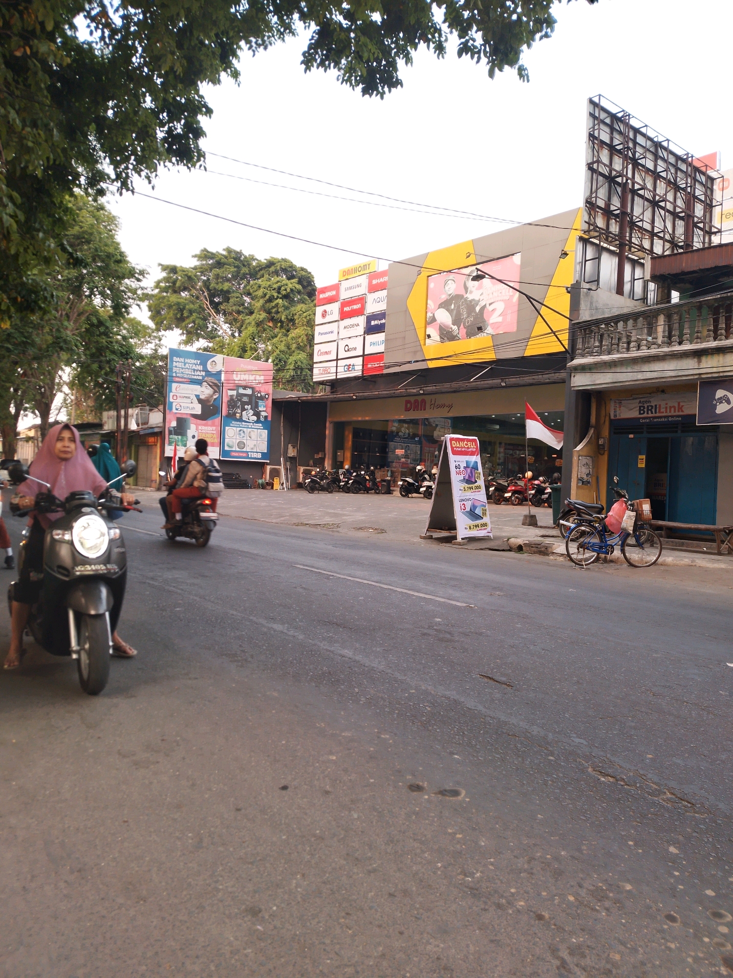 The image depicts a street scene in Kertosono, Indonesia, with a focus on a commercial building and passing traffic. In the foreground, a person on a dark-colored scooter is seen from the side, wearing a pink headscarf. The scooter has a license plate visible, reading "AG 3405 WS". Further down the street, another scooter carrying two individuals is passing by. The commercial building features a large yellow and grey facade with prominent signage, including "DAN Homy" and advertisements for various brands like LG, Samsung, and Polytron. A large billboard with an image of a couple is also present. In front of the building, a red and white Indonesian flag is displayed. Several motorcycles are parked in front of the building. To the right, a blue bicycle with a basket containing a red bag is parked on the sidewalk. A BRI Link agent office is also visible. The street itself is paved and shows signs of wear, with some water or oil stains. Trees line the left side of the street, and the sky is overcast.