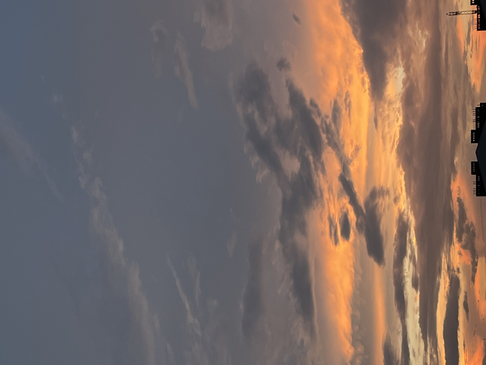 This is a vertical, medium-shot photograph taken at an upward angle of a sky filled with clouds during a sunset. The sky transitions from a dark gray at the top to vibrant orange and yellow hues closer to the horizon, indicative of sunset. There are several buildings visible at the bottom of the image, appearing silhouetted against the bright sky. One building on the far right has a tall, thin structure attached to it that resembles a construction crane. The image does not contain any people or animals. The scene depicts an urban landscape at dusk.