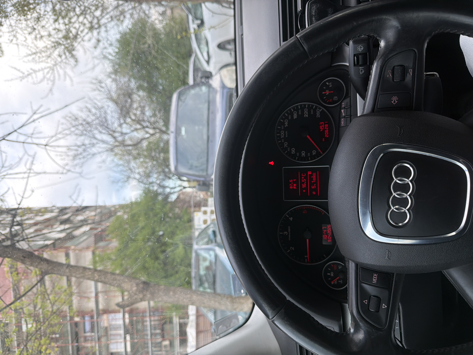 The image shows the interior of an Audi car, focusing on the steering wheel and dashboard. The dashboard displays information such as the radio station (87.5 FM1), temperature (16.5°C), fuel consumption (5.1 l/100km), time (18:47), date (9.04.2026), speed (49.3 km/h), and mileage (206281 km). Outside the car, the scene is blurred but suggests an urban environment with trees, parked cars, and buildings. Based on the blurred buildings and parked cars visible through the windshield, it is difficult to pinpoint the exact location within Plovdiv, Bulgaria. However, the presence of trees and buildings indicates a residential or urban street.