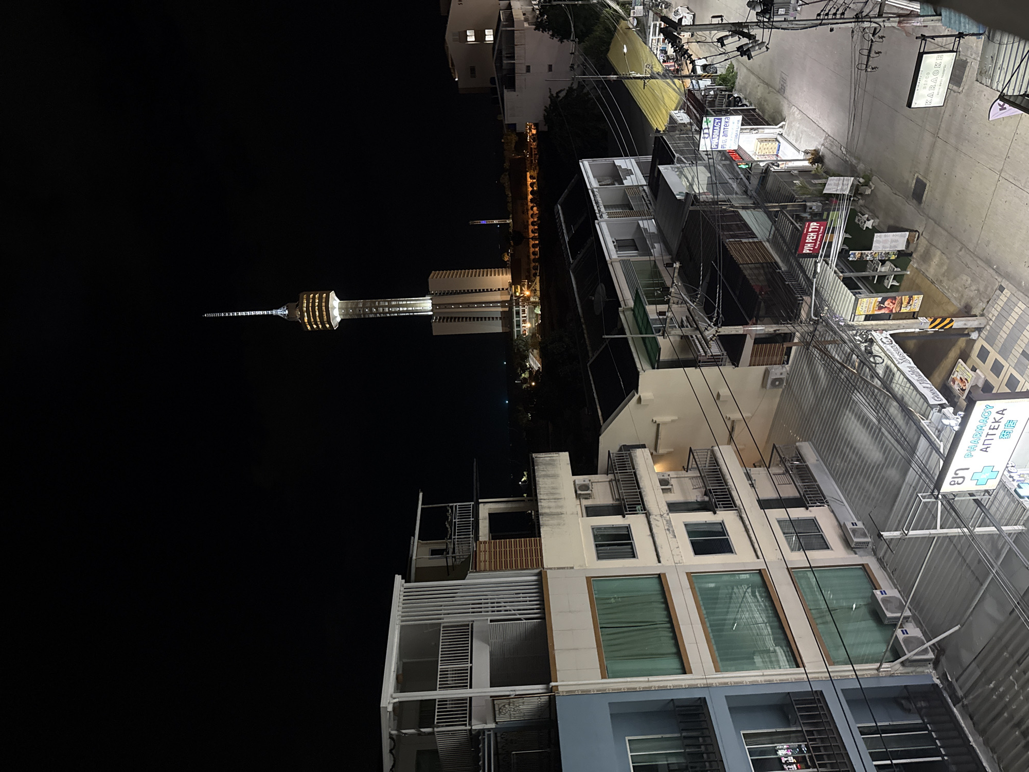 This is an eye-level, wide shot of a city street at night in Pattaya City, Thailand. The scene is dominated by a tall, brightly lit skyscraper with a unique, tiered top and antenna. In the foreground, there are several multi-story buildings with balconies and windows, some of which are illuminated. Below, the street is lined with small businesses, including a pharmacy with a green cross logo, a karaoke sign, and various other shops with signage in what appears to be Thai and possibly other languages. Power lines crisscross the scene, and there are a few parked cars visible on the street. The overall impression is one of a bustling urban environment at night.