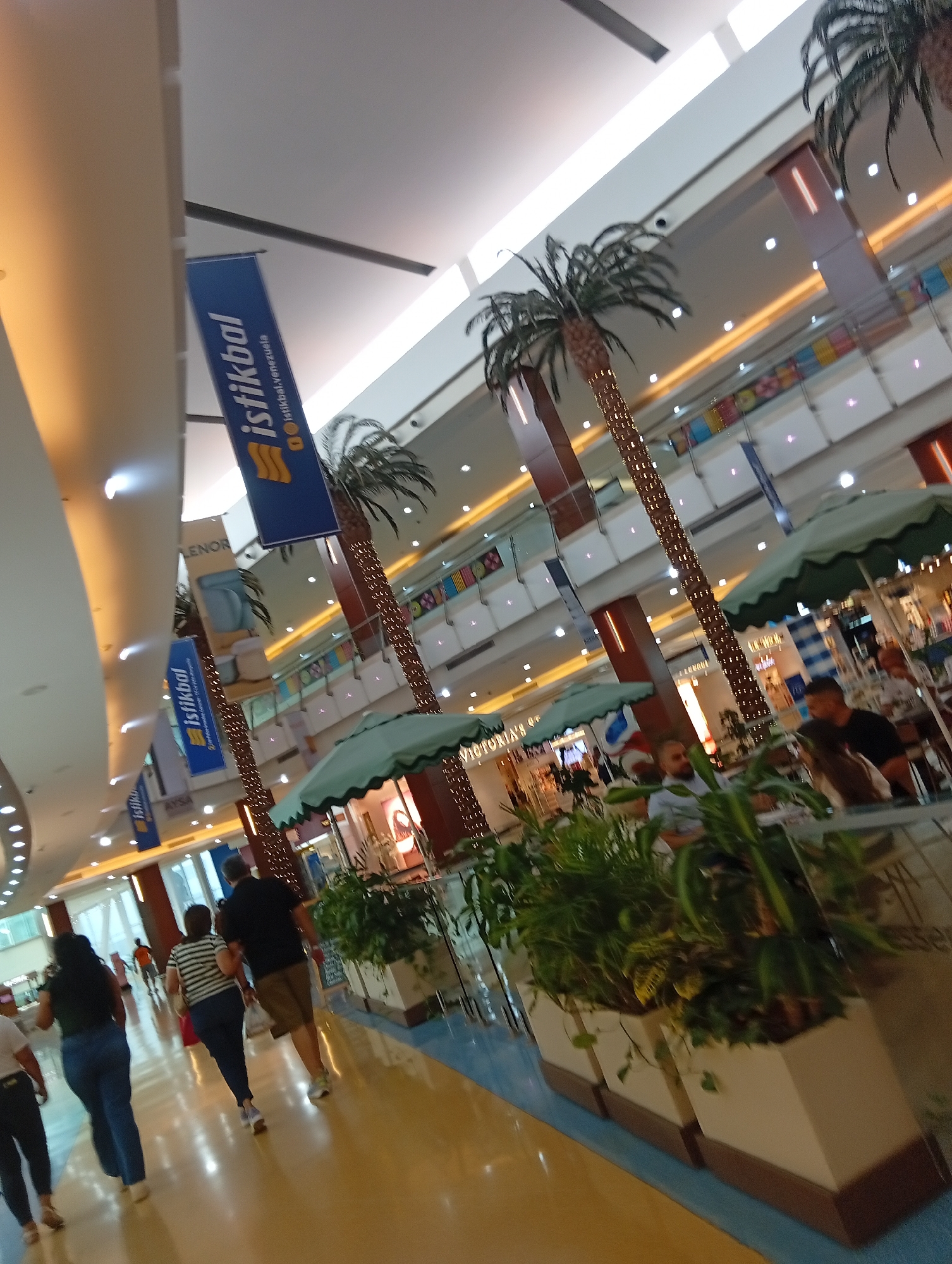 The image shows the interior of the Sambil Margarita shopping mall in Porlamar, Venezuela. Several people are walking along a brightly lit, yellow-toned floor. Two women in casual clothing, one with a black top and the other with a striped top, walk away from the camera. A man in a black shirt and khaki shorts walks near them. The mall is multi-level, with palm trees adorned with lights reaching towards the ceiling. Banners with the "Istikbal" logo hang overhead. Plants in large white planters are positioned near cafe tables and umbrellas. People are sitting and eating at the cafe tables. Several store fronts are visible in the background.
