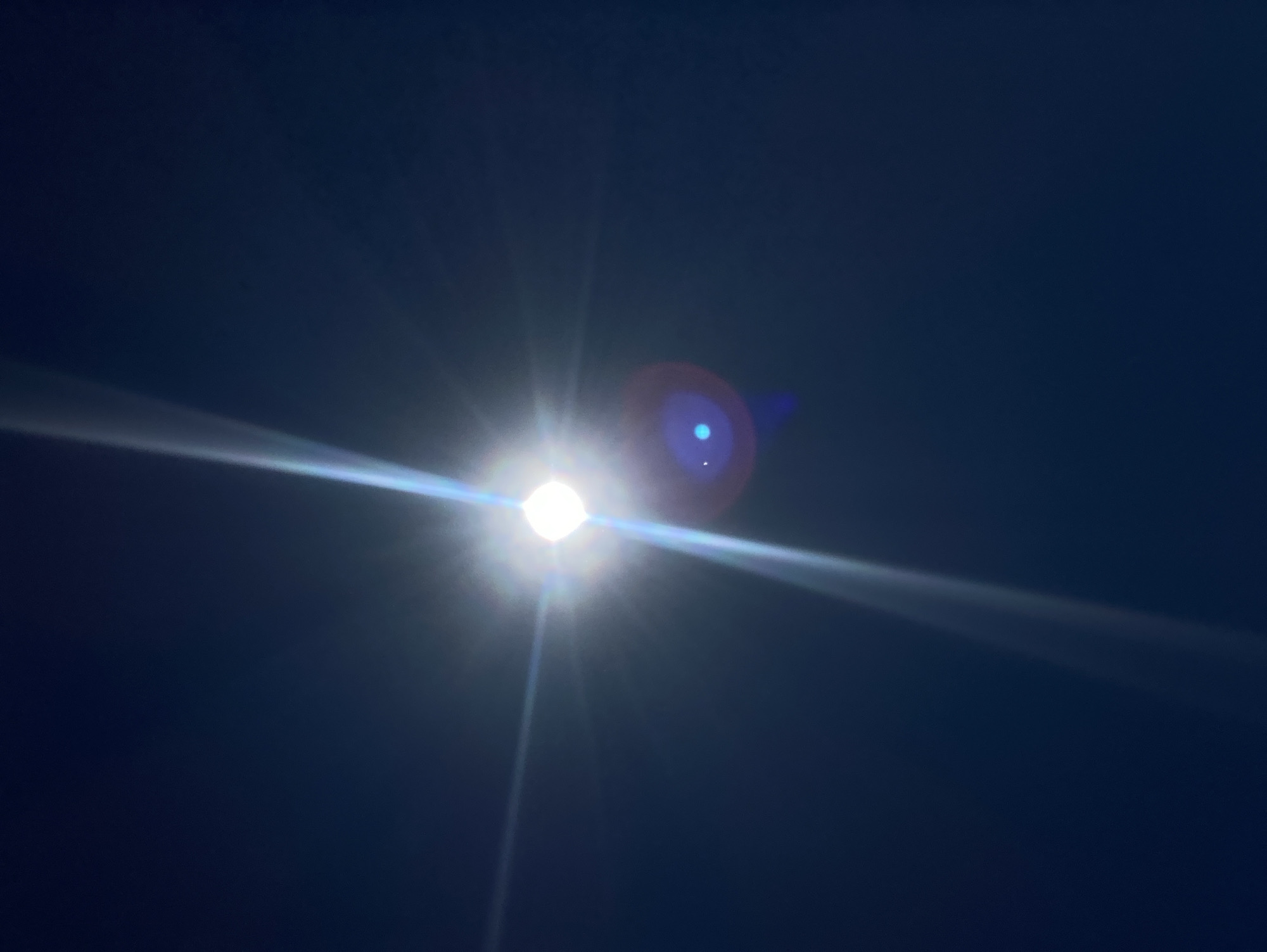 The image depicts the sun as a bright, luminous sphere against a dark blue sky. Prominent lens flares emanate from the sun, creating streaks of light that extend outwards. One particularly strong flare, a thin white beam, stretches diagonally from the lower left to the upper right corner of the frame. Another, more diffuse flare, displays a circular shape with a blueish tint and a smaller, brighter blue dot within it, positioned to the lower right of the sun. The sky itself is a deep, uniform blue, devoid of any visible clouds or celestial bodies other than the sun. The setting is outdoors, under an open sky. There are no people, animals, or man-made structures visible in the image. The image's metadata indicates it was taken in Celo Rakity, Russia.