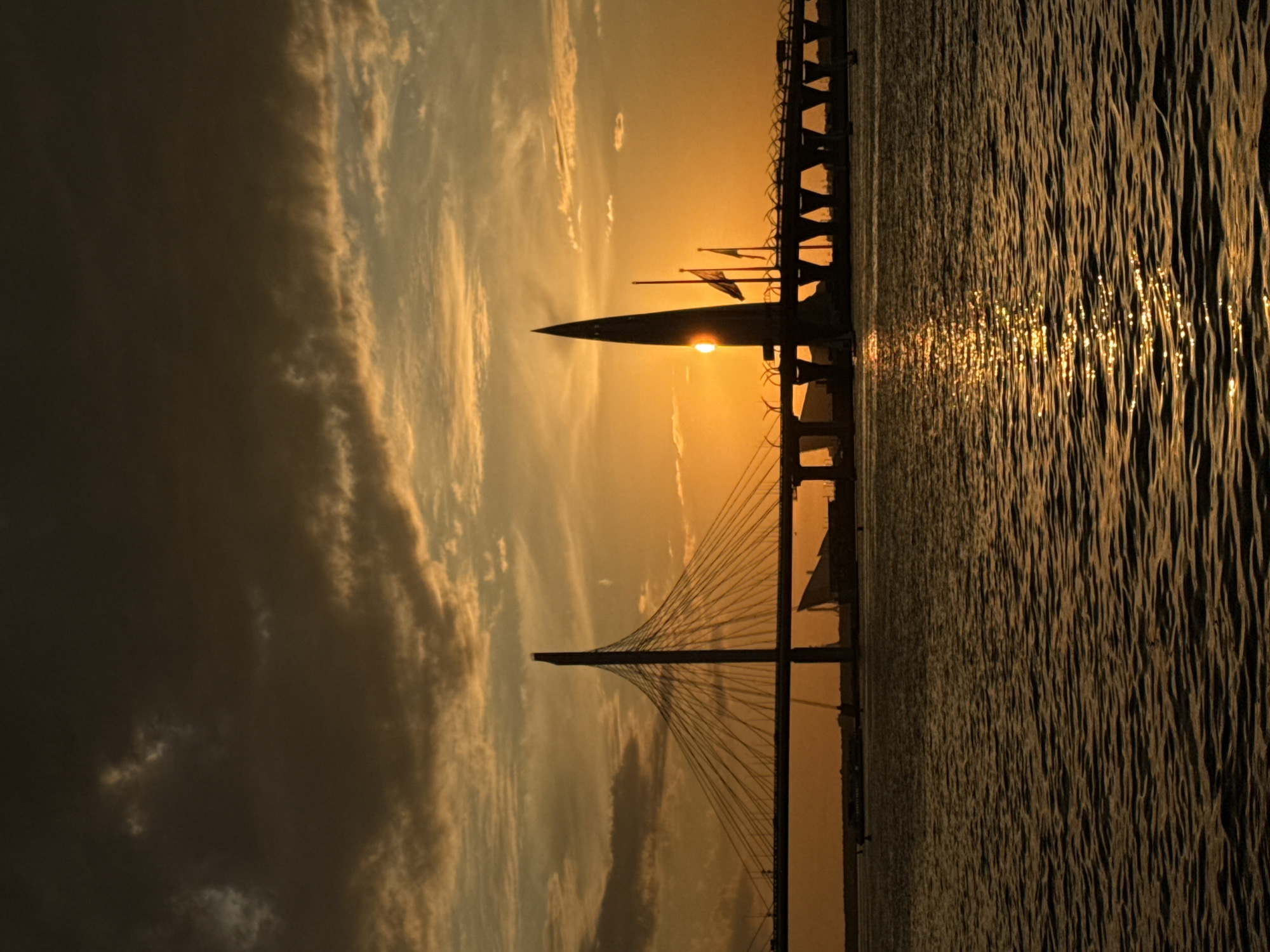 This media file captures a stunning sunset or sunrise over a large body of water, presenting a dramatic silhouette scene. The central subjects are a majestic cable-stayed bridge and a prominent, pointed silhouette, likely a large ship or monument, perfectly positioned to obscure the sun.
The sky is bathed in rich golden and orange hues around the horizon, transitioning to softer yellows and then darker tones further away from the sun. Scattered clouds are present, with more substantial, darker cloud formations visible on the left side of the frame, adding depth to the sky.
The water surface in the foreground and midground is highly reflective, shimmering with bright, elongated streaks of golden light that catch the low sun, indicating gentle ripples. On the far right, another bridge structure or pier is partially visible, also in silhouette. The sun's position directly behind the pointed silhouette creates a striking halo or sunburst effect around it, enhancing the dramatic light play.
The scene is devoid of human activity, focusing on the interplay of natural light and man-made structures. The warm, luminous colors strongly suggest the time of day is either dawn or dusk. Given the location context of Saint Petersburg, Russia, the cable-stayed bridge is likely a component of the Western High-Speed Diameter, and the distinctive pointed silhouette could represent a notable vessel or a landmark along the Neva River or Gulf of Finland. There is no visible text in the image.