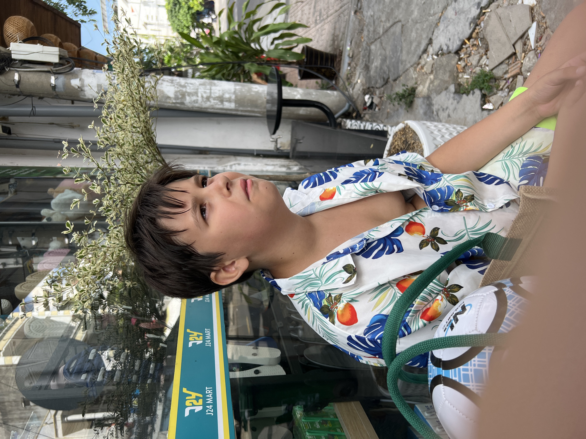 The media file is a portrait of a young boy, viewed from the shoulders up, outdoors. He has dark hair and is looking to his left with a neutral expression. He is wearing a short-sleeved, open-necked shirt with a vibrant tropical print featuring large blue leaves and orange fruits. The boy is positioned in front of a shop window with a yellow and teal sign that reads "J24 MART".
Behind the boy, a leafy green plant with small white and green leaves is visible, partially obscuring the background. Further back, there is a concrete sidewalk with some scattered debris. The overall setting appears to be a street scene, likely in a commercial area, given the shop sign and the outdoor cafe-style seating visible in the background on the right. The lighting suggests it is daytime, possibly sunny, as there are bright highlights. The image is cropped closely on the boy, with a large portion of the right side of the frame out of focus and blurred, suggesting a shallow depth of field. A green rope and the edge of a bag, along with a white and blue soccer ball, are visible in the lower right quadrant, close to the camera. The content and tropical print suggest a warm climate, consistent with the provided location context of Nha Trang, Vietnam.