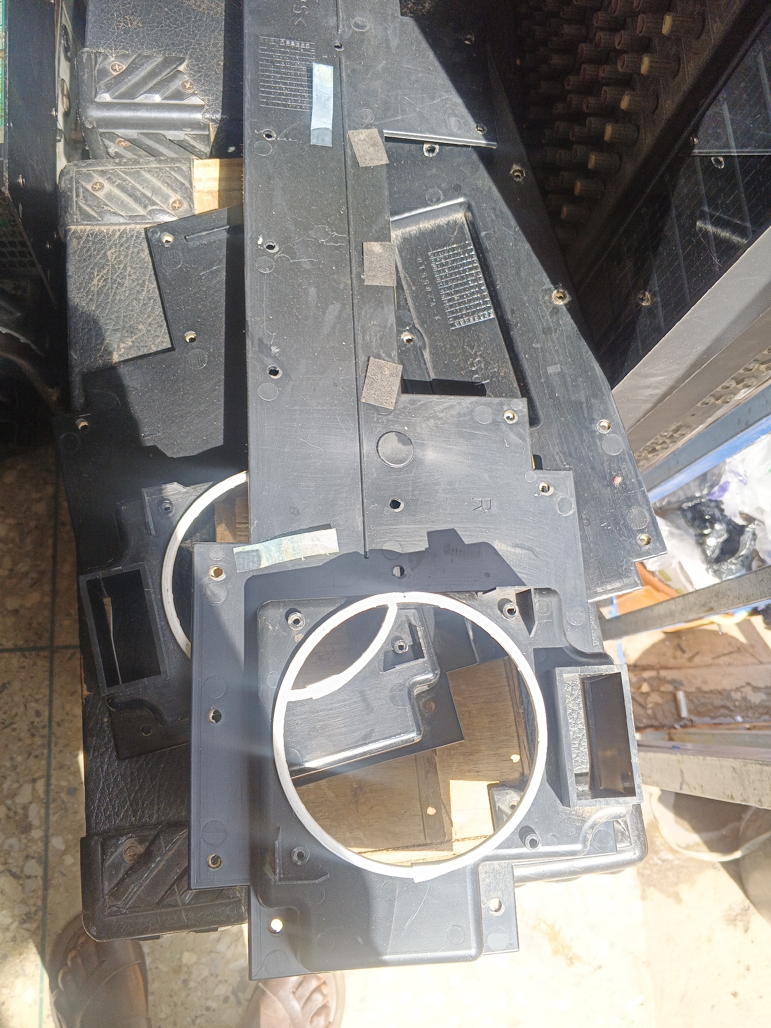 The image depicts a close-up, overhead view of disassembled black plastic components, possibly from an electronic device, resting on a tiled surface. The components are layered and appear dirty, with visible screws and mounting points. Some of the plastic pieces have textured surfaces. In the background, there is a hint of a wooden surface and a blurred area suggesting a more cluttered environment with plastic bags. A person's bare feet, wearing sandals, are visible in the lower left corner of the frame, indicating their presence at the scene. The lighting suggests an outdoor or well-lit indoor environment, casting shadows on the objects. The scene is forensic in its depiction of the disassembled parts, presenting them without context or judgment.