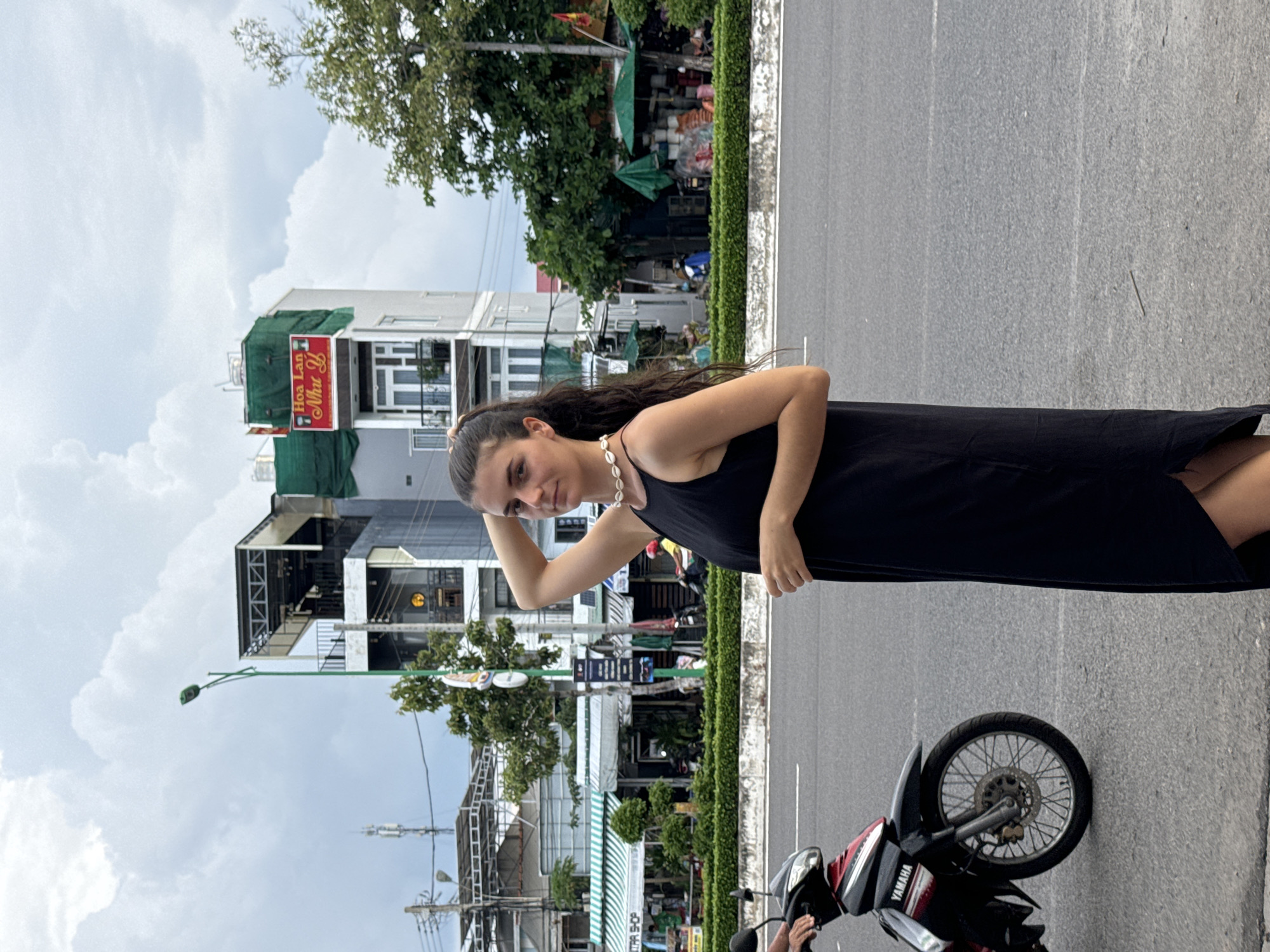 A young woman with long, dark hair stands on the edge of a paved road, posing for the camera. She wears a black sleeveless dress and a white, shell-like necklace. Her right arm is bent with her hand near her head, and she has a calm expression, looking directly at the viewer. To her right, a red and black Yamaha scooter is parked, with a matching helmet resting on its handlebars.
The scene depicts an outdoor street environment in Phan Thiết, Vietnam. The woman is standing next to a narrow strip of well-maintained green bushes, which separates the road from the buildings beyond. In the midground and background, there are multi-story buildings of varying architectural styles, suggesting a mix of commercial and residential spaces. One building prominently features a red and yellow sign with Vietnamese text, "Hoa Lan" and "Như Ý". Another building has a green tarp covering part of its exterior, and further down, a shop with a green and white striped awning is visible. Utility poles, wires, and streetlights are scattered throughout the background. The sky is overcast with visible clouds, indicating daytime. The general ambiance is relaxed, typical of a casual street scene.